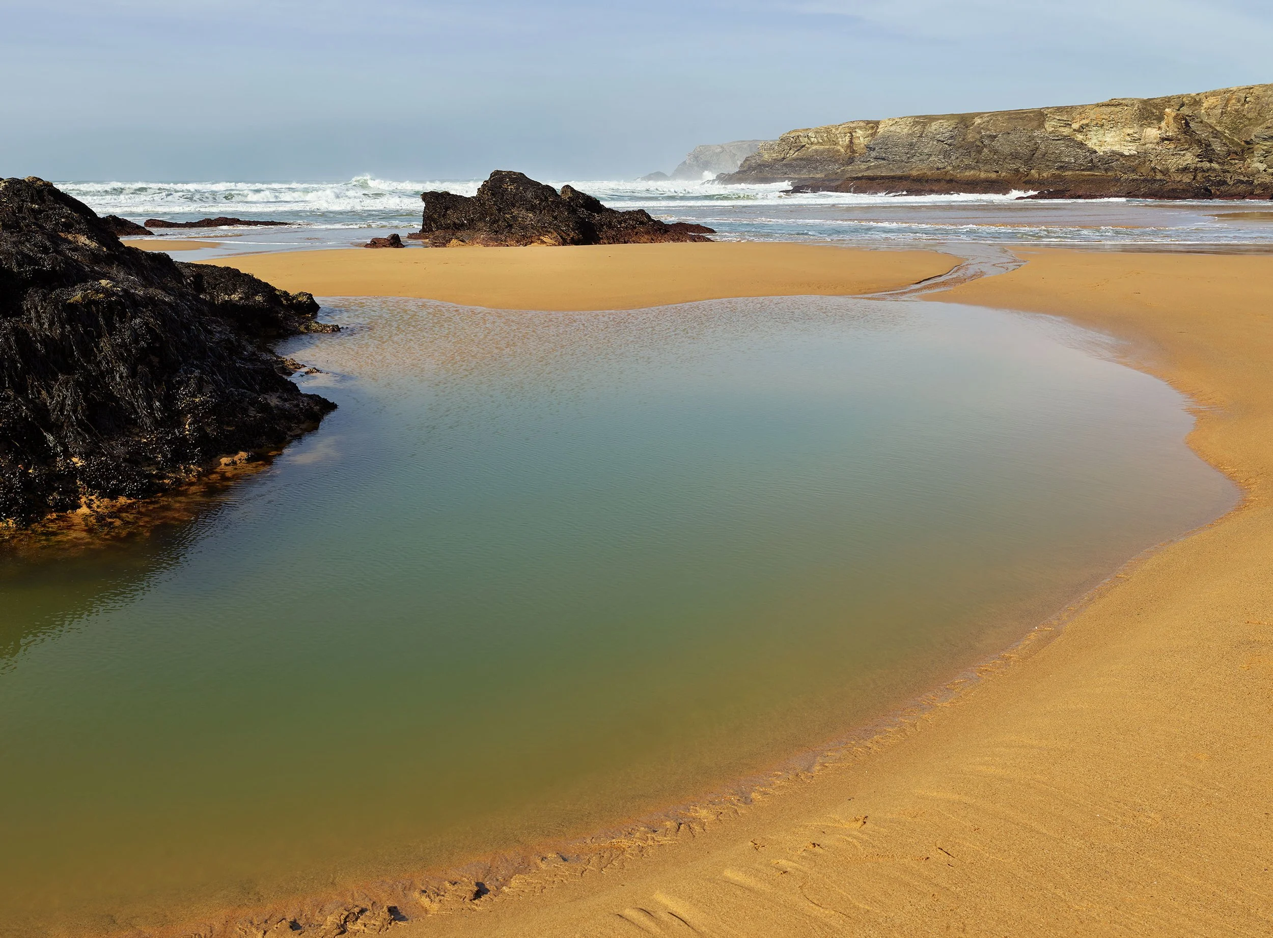 Plage de sable doré avec des rochers noirs, des vagues qui déferlent en arrière-plan et des falaises en hauteur au loin, ciel partiellement nuageux.