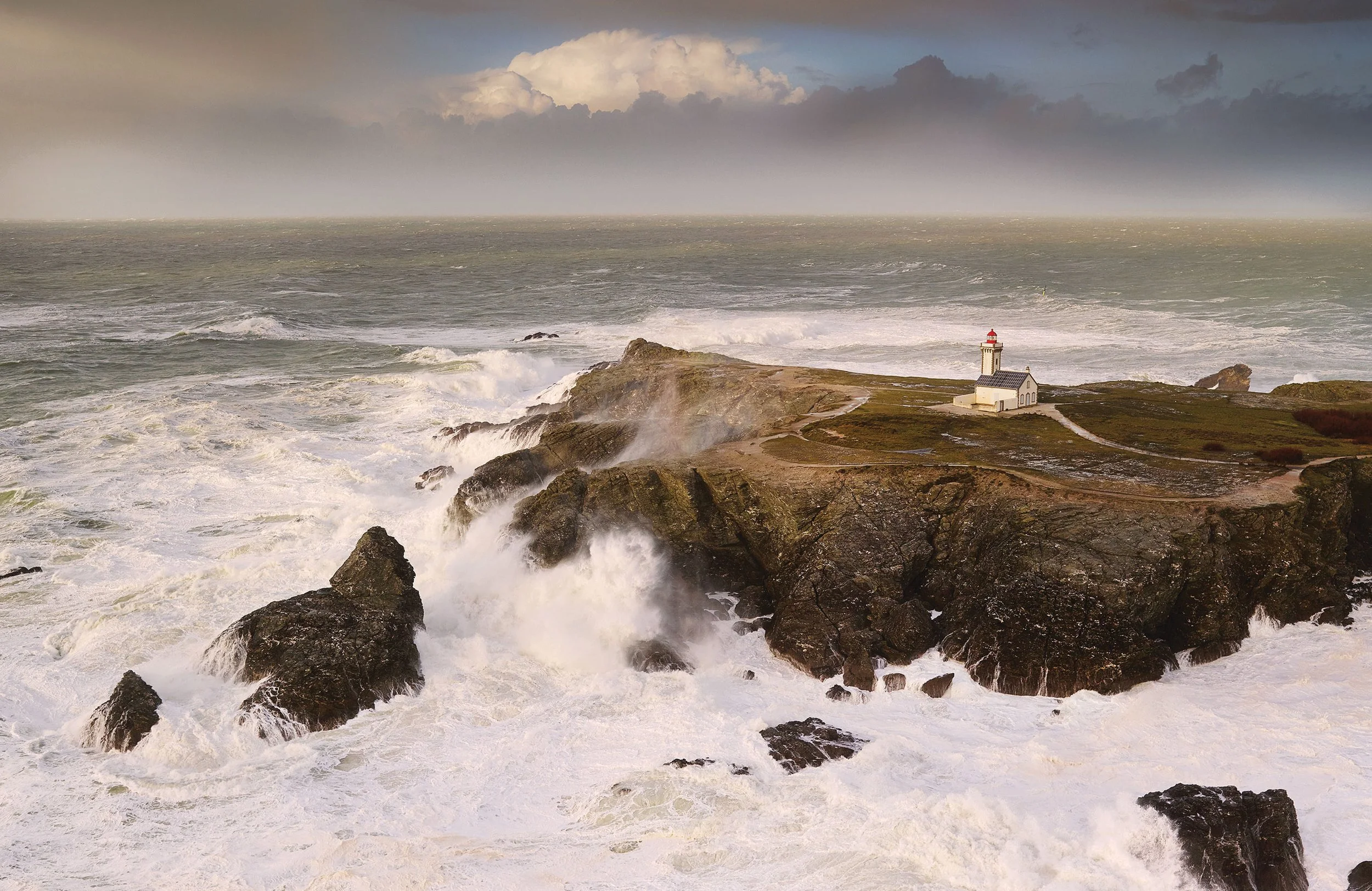 Une scène maritime avec des vagues déferlantes contre des rochers, un phare blanc avec un toit rouge sur un promontoire rocheux, sous un ciel nuageux avec des éclairs
