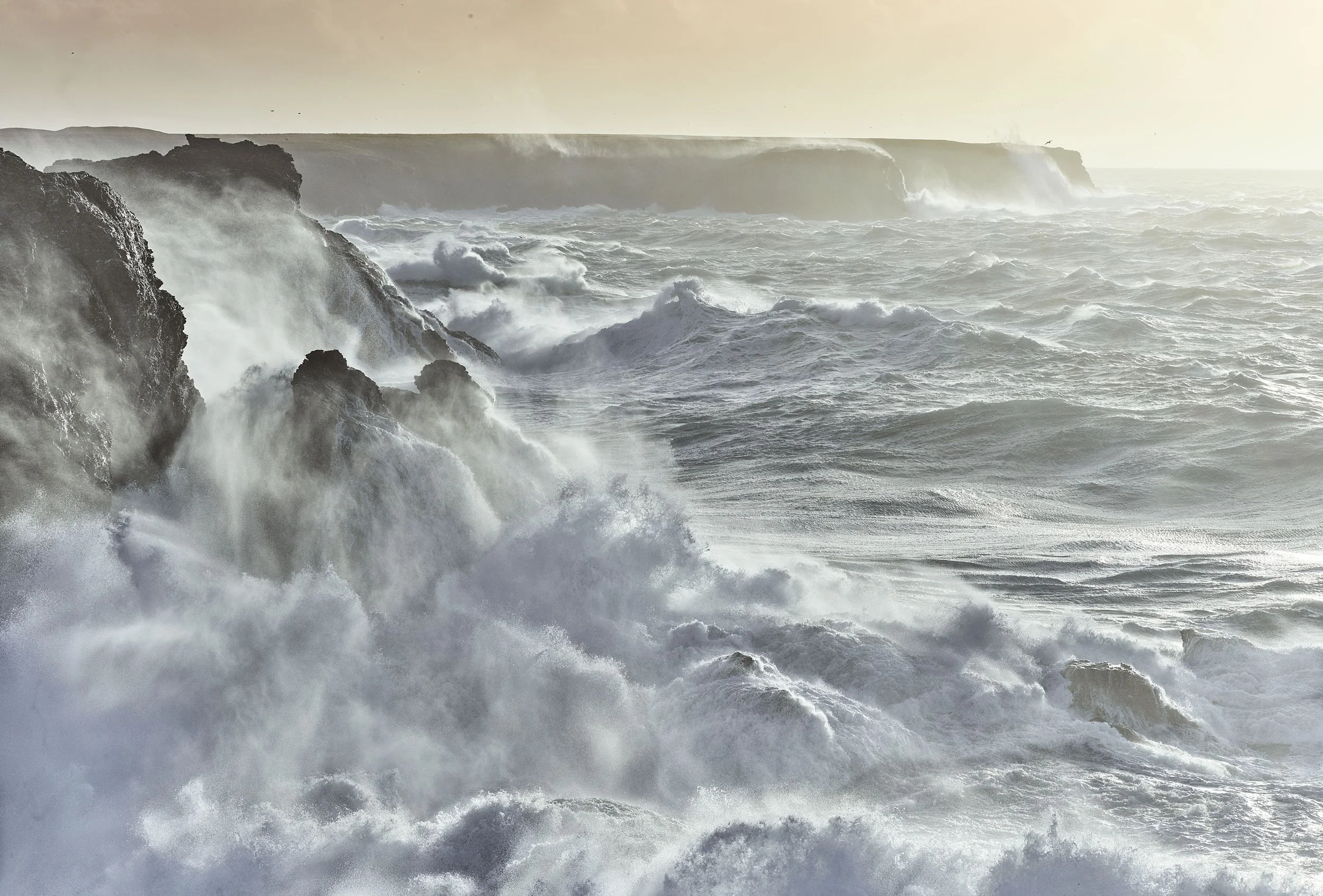 Vagues déchaînées s'écrasant contre des falaises rocheuses au bord de l'océan, sous un ciel brumeux et lumineux.