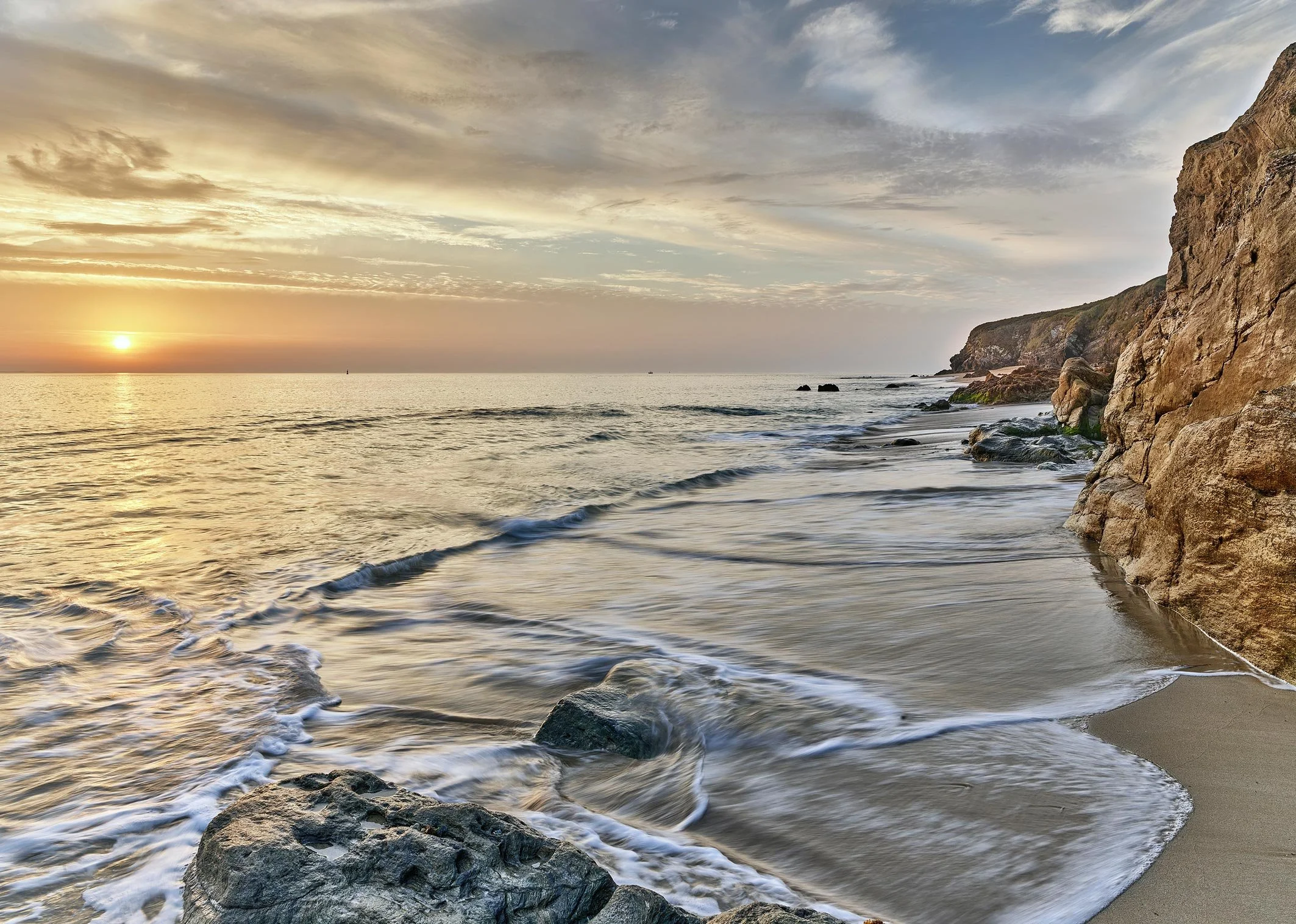 Coucher de soleil sur une mer calme avec des rochers au premier plan et une falaise sur la droite. Le ciel est partiellement nuageux avec des couleurs chaudes du soleil couchant.