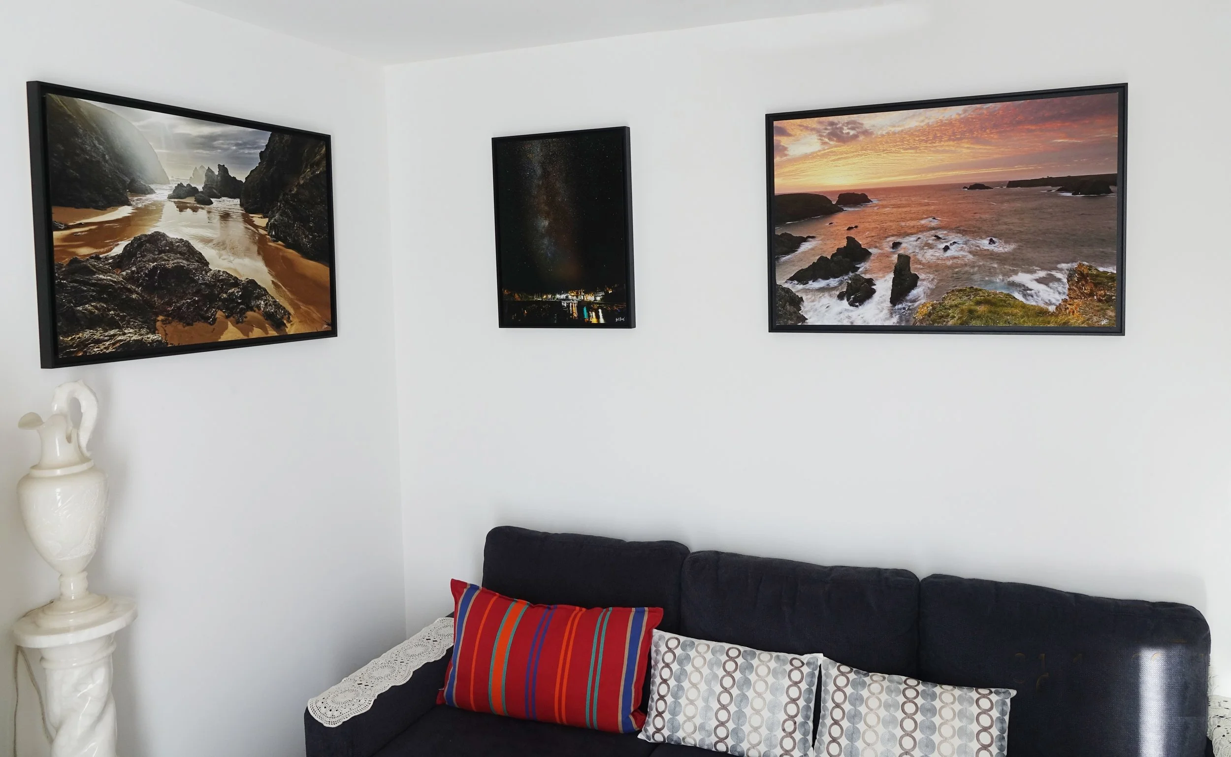 Salle d'exposition avec trois tableaux de paysages maritimes et une lampe blanche sur un mur blanc, un canapé noir avec des coussins colorés.
