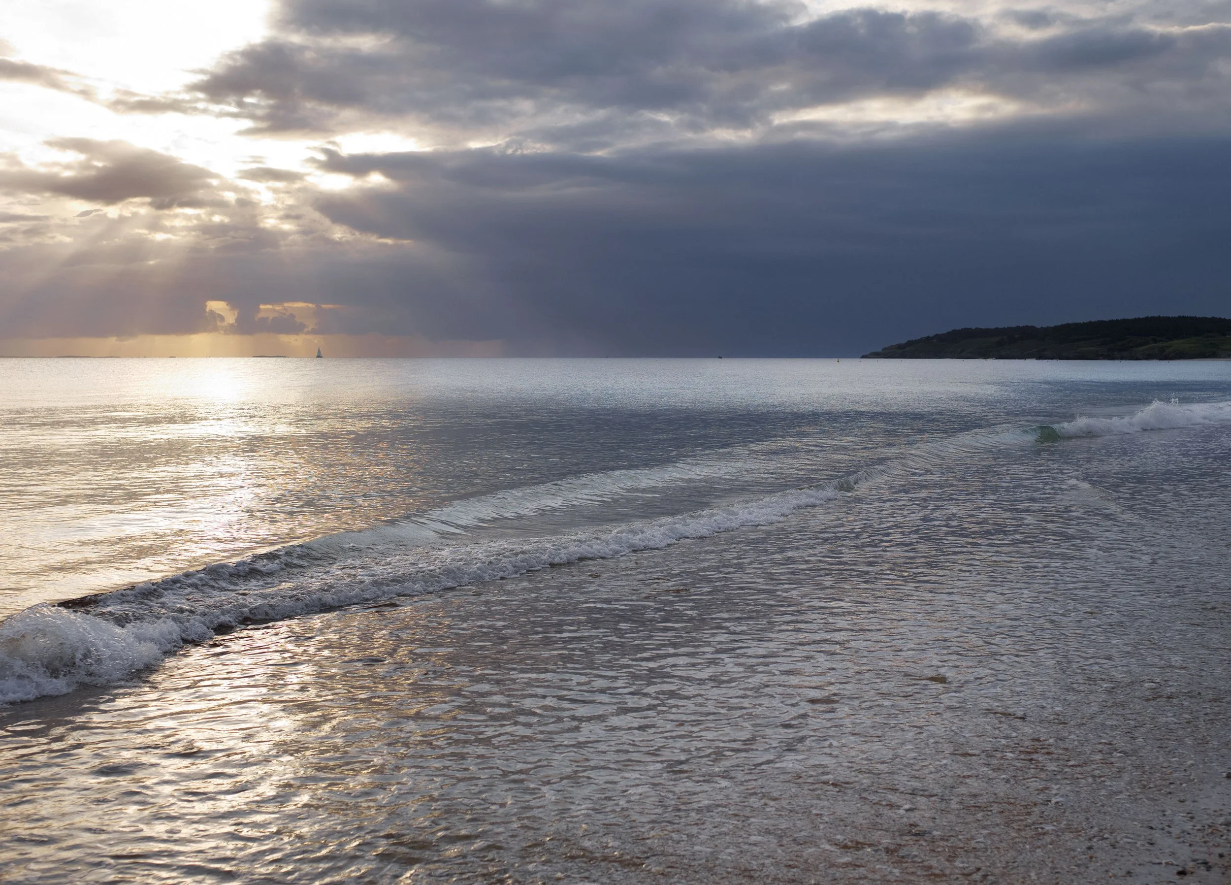 Coucher de soleil sur la mer avec des nuages et un voilier en arrière-plan, vue depuis une plage avec des vagues
