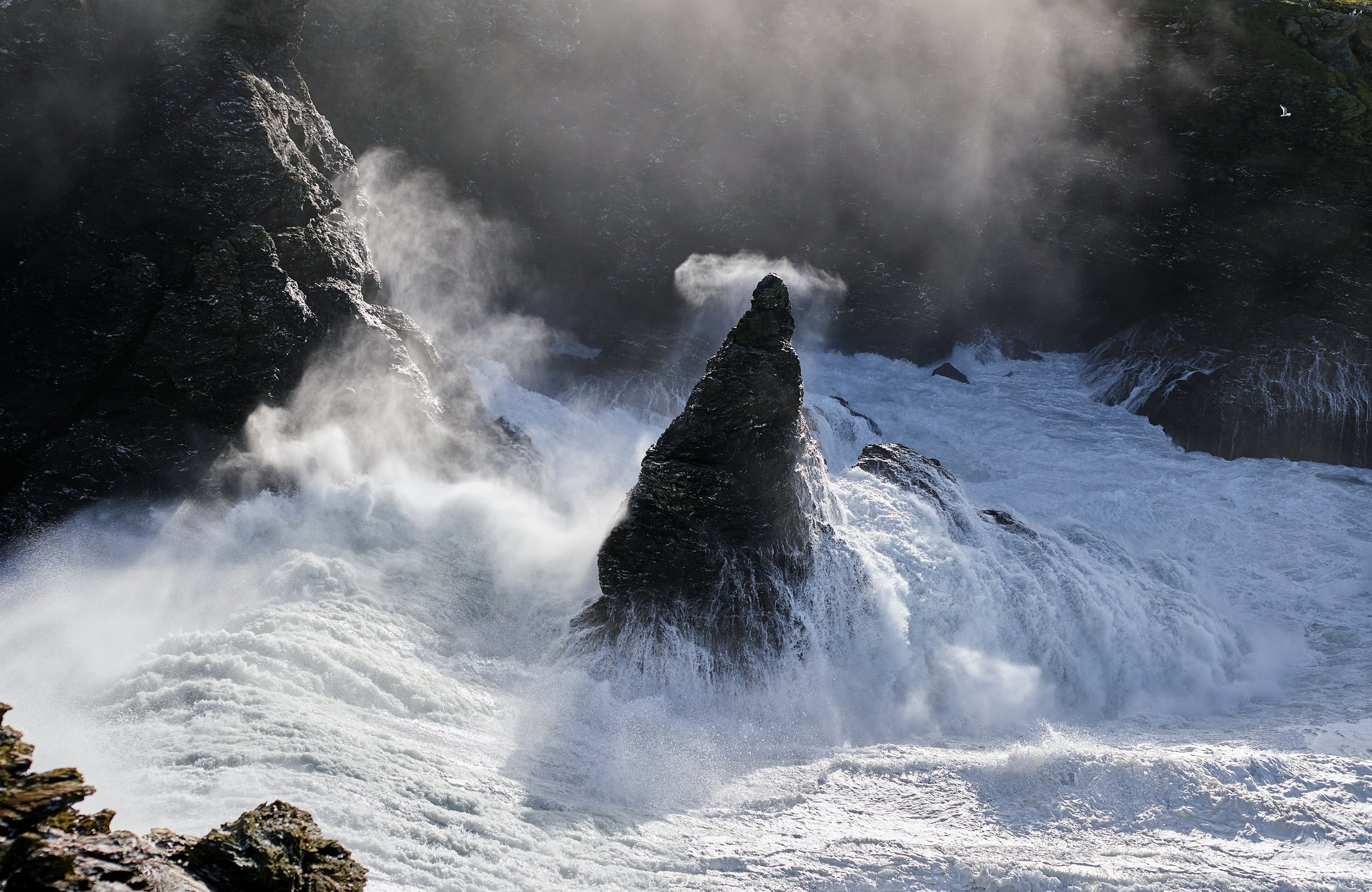 Vagues déferlantes contre des rochers escarpés dans une mer agitée, avec de la vapeur ou de la brume en arrière-plan.