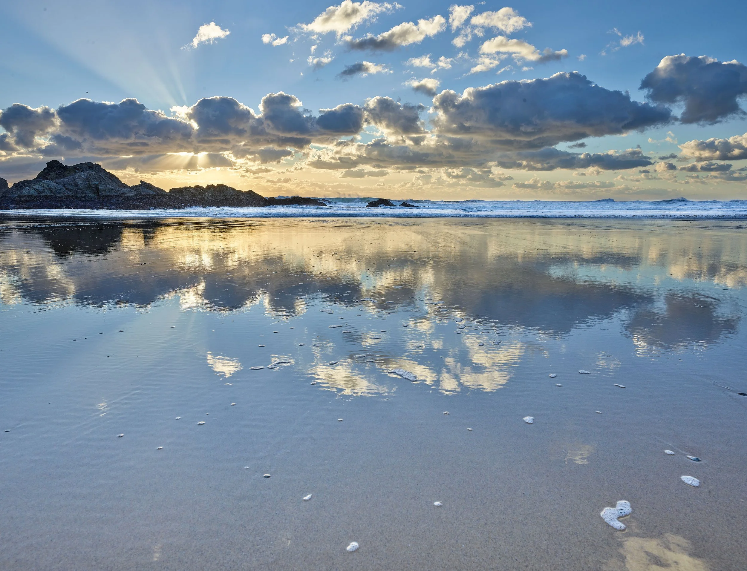 Plage avec des rochers, des vagues et un ciel nuageux avec le soleil couchant.