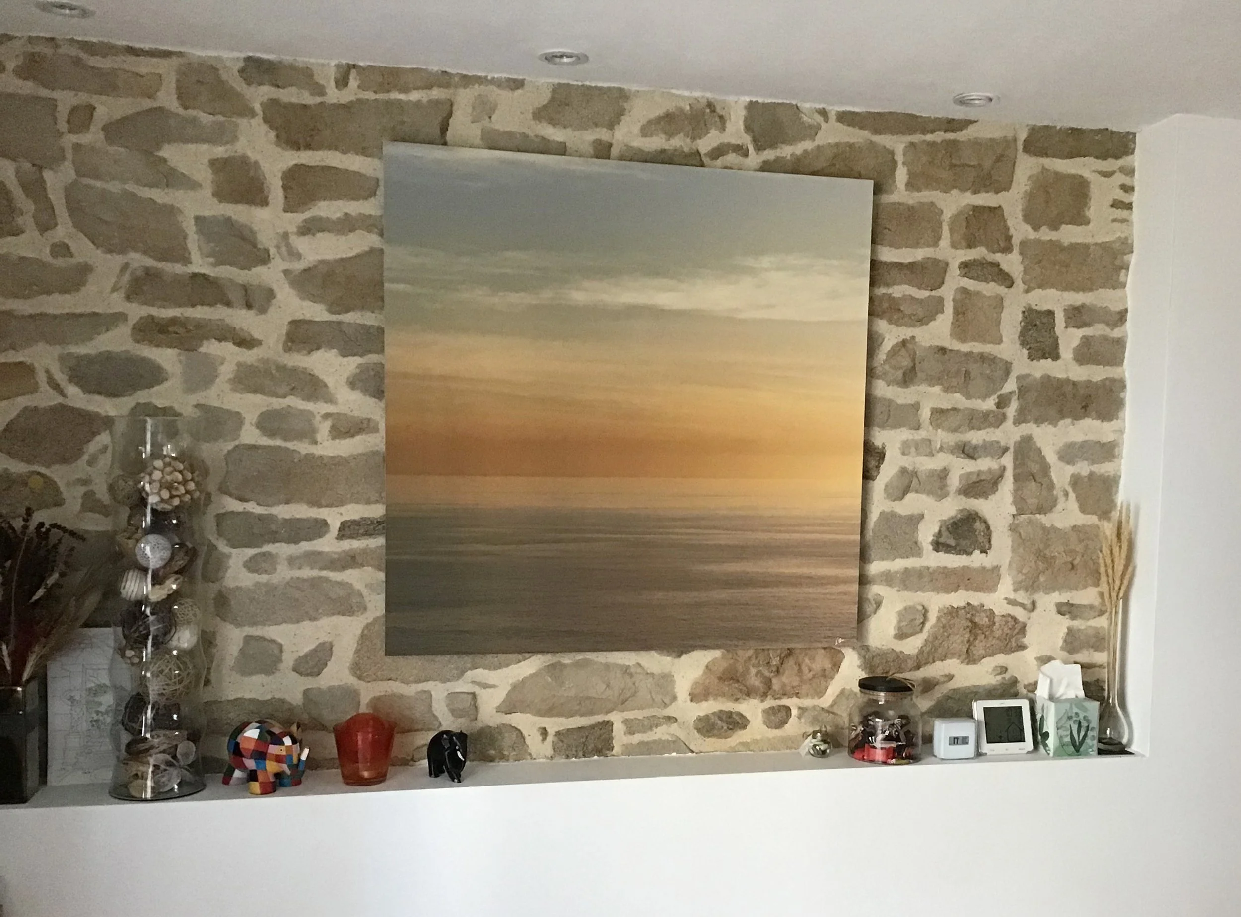 Décor intérieur avec un mur en pierre, une peinture abstraite sur le mur, une étagère avec des objets décoratifs tels qu'une sphère colorée, un vase en verre orange, des figurines en bois, un thermomètre numérique, et une boîte à mouchoirs.