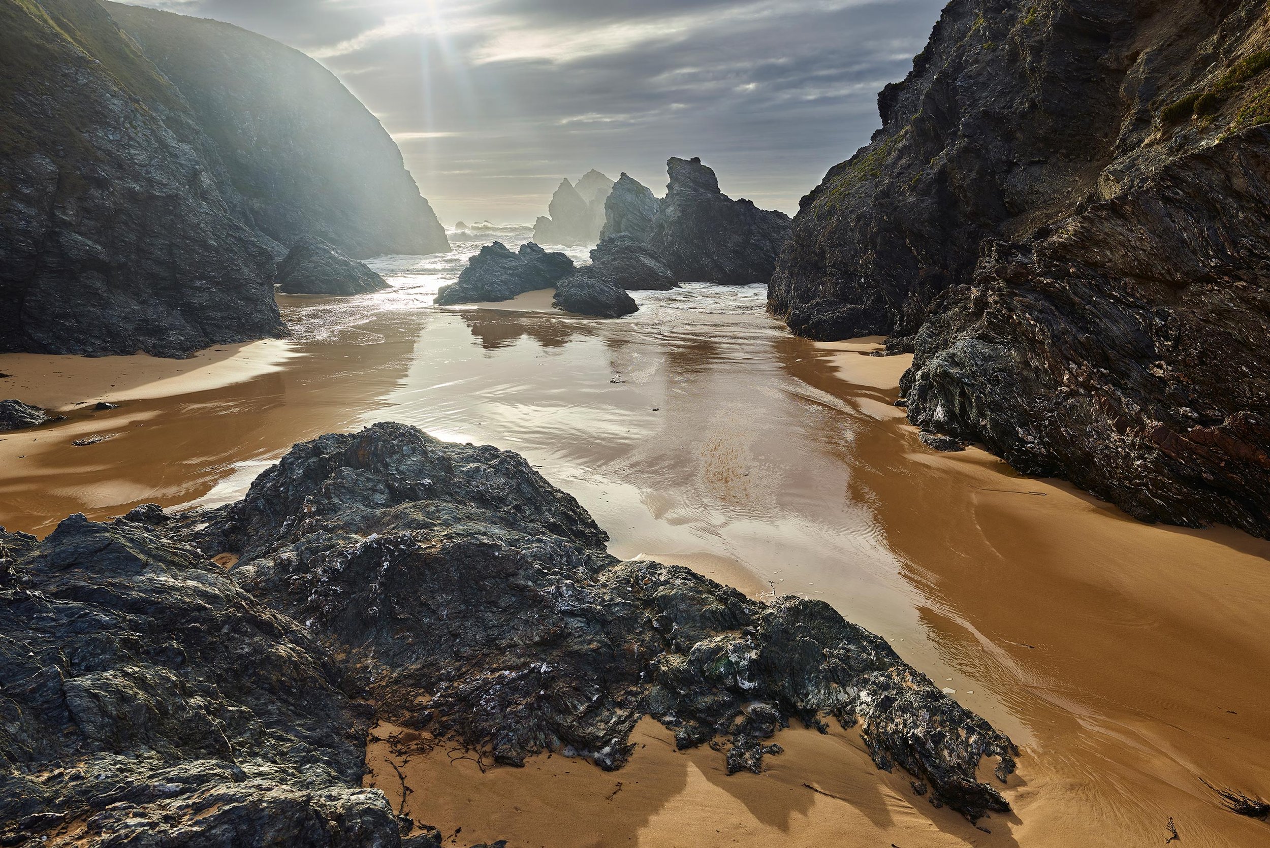 Côte rocheuse avec des rochers noirs sur une plage de sable doré, vagues de mer, ciel nuageux, lumière du soleil perçant à travers les nuages.