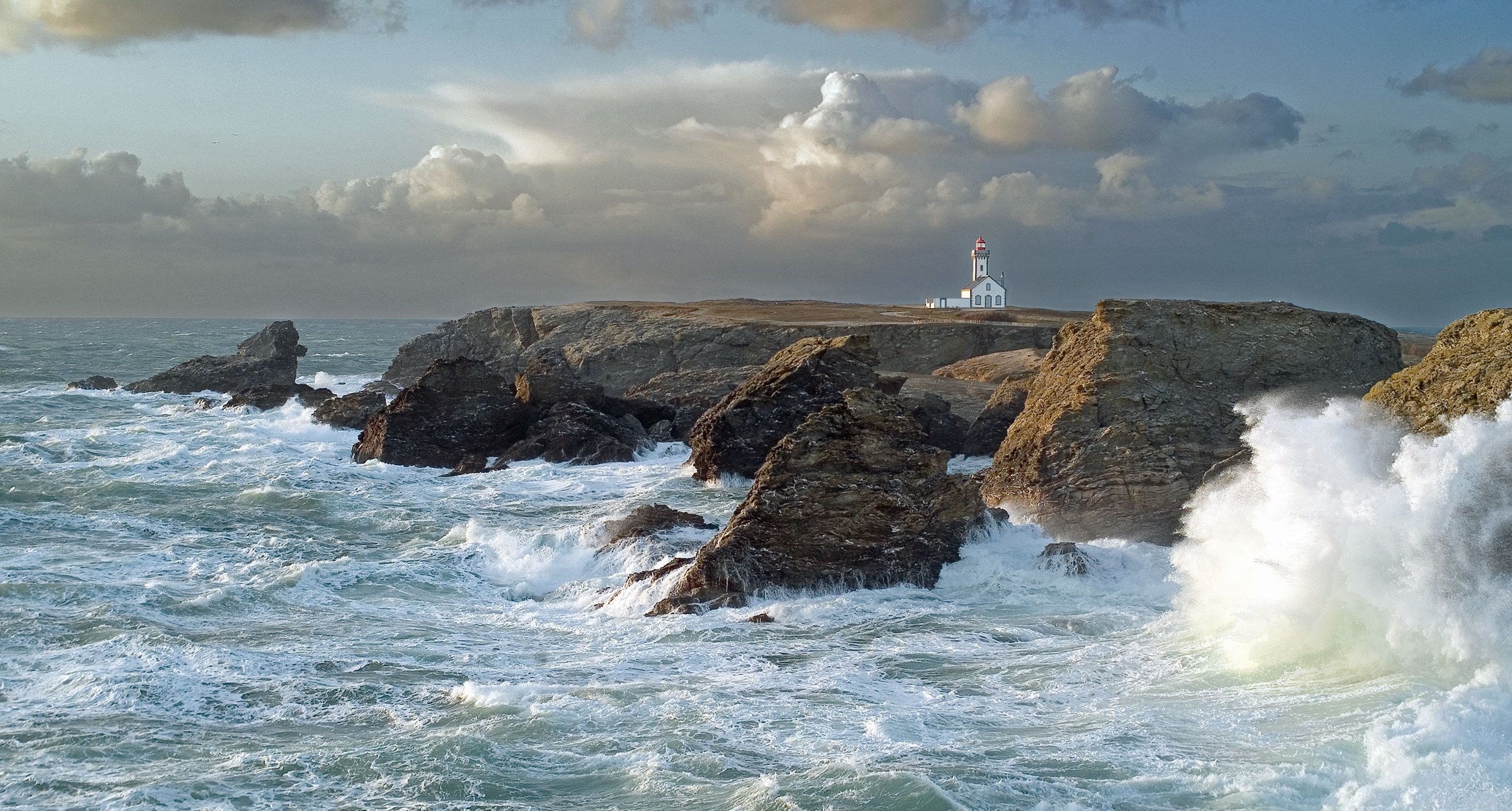 Un phare blanc situé en haut de falaises rocheuses bordant la mer, avec des vagues s'écrasant contre les rochers et un ciel nuageux.