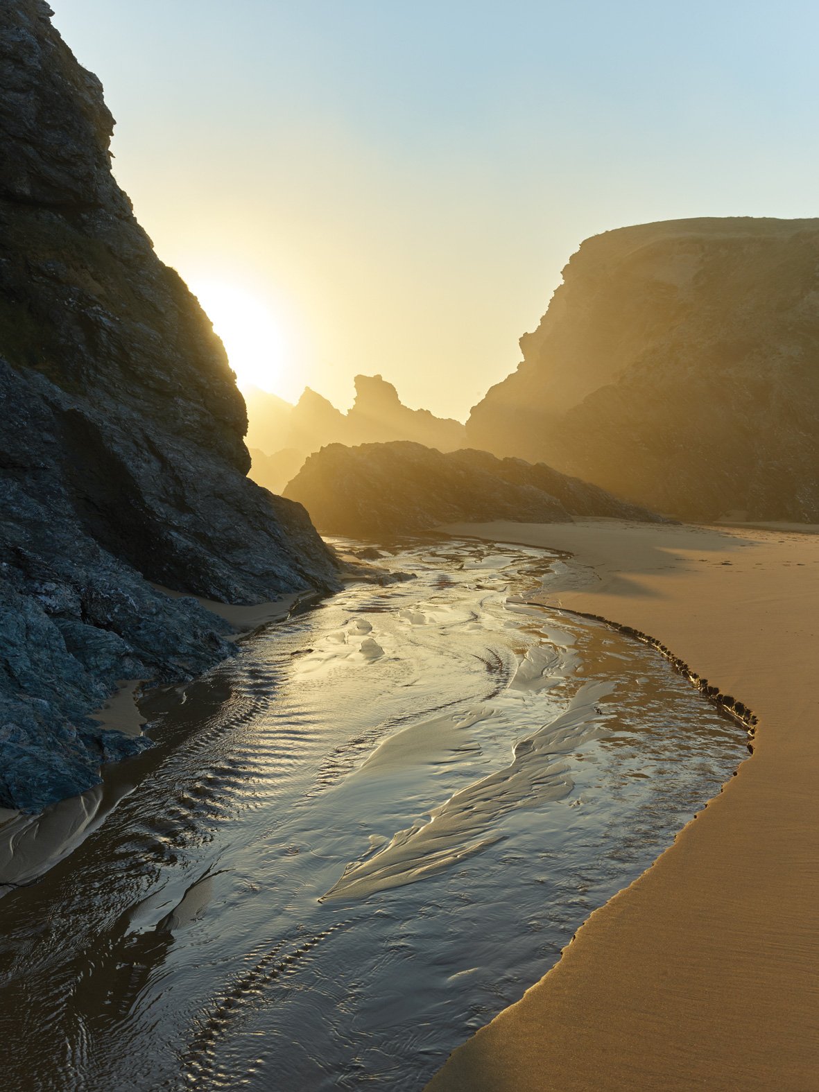 Côte rocheuse avec une petite rivière ou un estuaire au coucher du soleil, entourée de falaises, plage de sable et lumière dorée