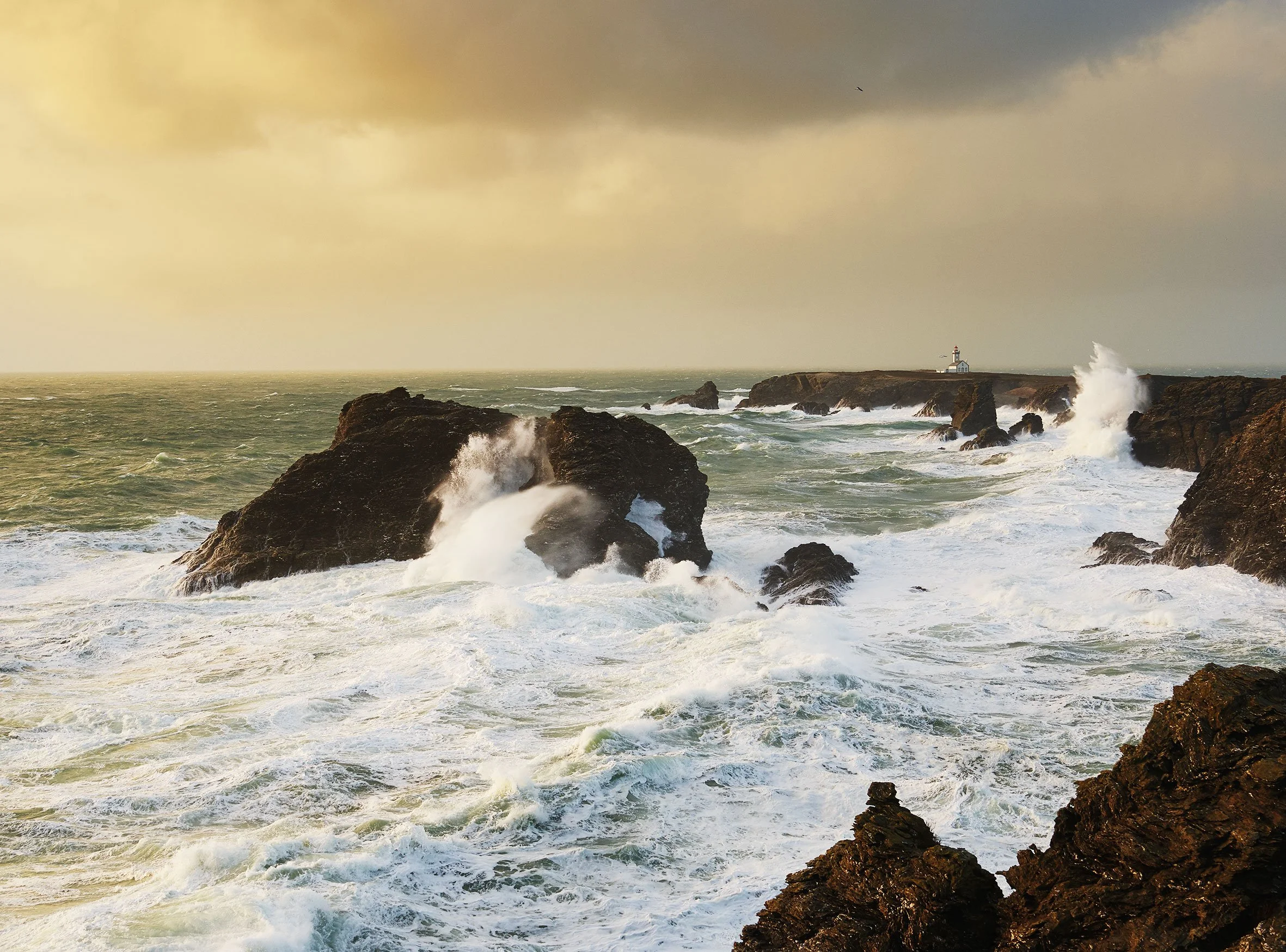 Mer agitée avec des vagues se brisant contre des rochers, un phare blanc sur la côte rocheuse au loin, ciel nuageux en arrière-plan.