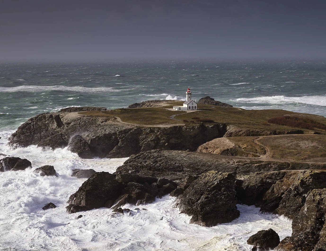 Phare blanc avec dôme rouge situé sur une falaise au bord de la mer, avec des vagues déferlantes et un ciel orageux en arrière-plan.