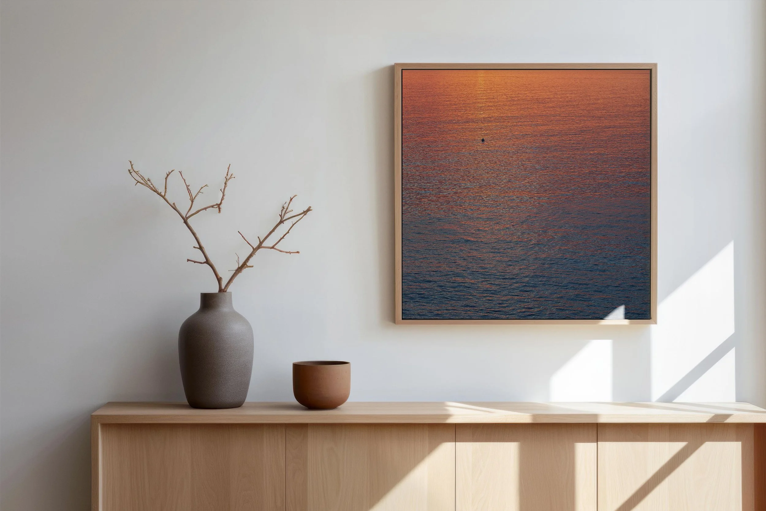 Une décoration intérieure minimaliste avec un tableau représentant une mer au coucher du soleil, une grande vase grise avec des branches et deux petits vases en terre cuite sur un meuble en bois clair.