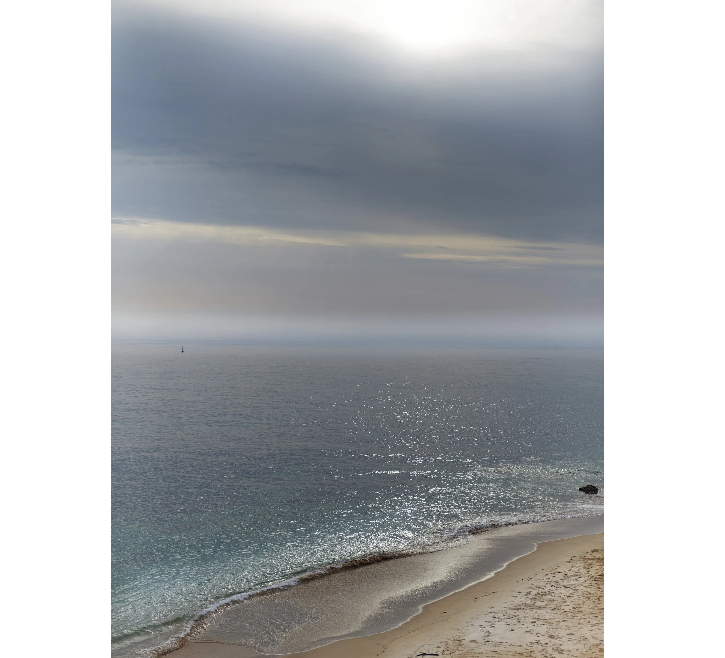Plage avec sable fin, mer calme et ciel nuageux, avec quelques rochers visibles en bas à droite.