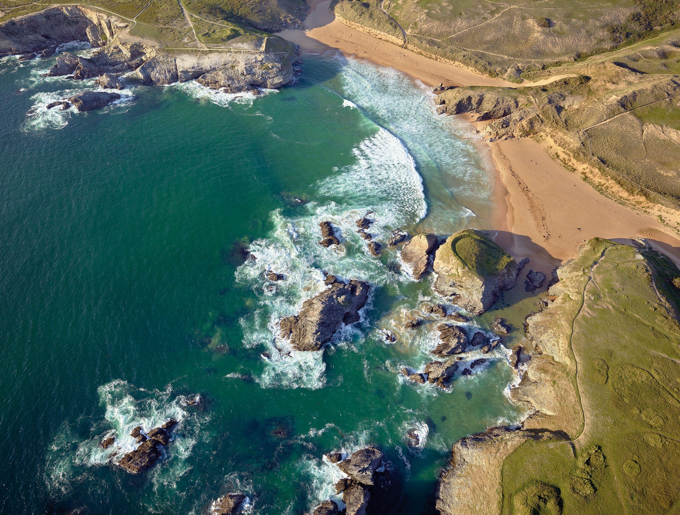 Vue aérienne d'une côte rocheuse avec une plage de sable beige et des eaux turquoise, entourée de collines verdoyantes.