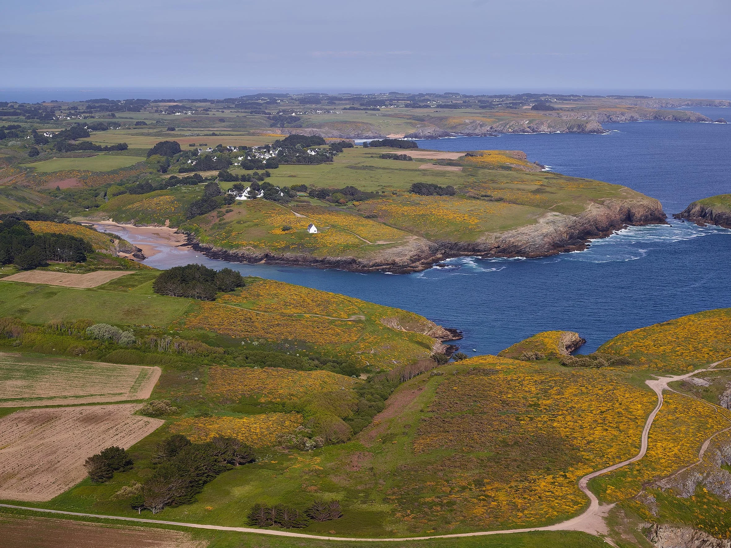 Paysage côtier avec des falaises, des plages, des champs verts, des maisons blanches et la mer au loin.