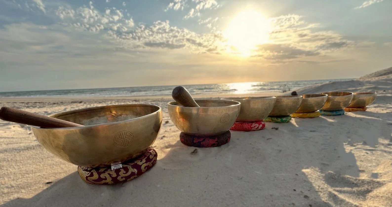 Seven Tibetan singing bowls placed on colorful cushions in a row on the beach with the ocean and sunset in the background.