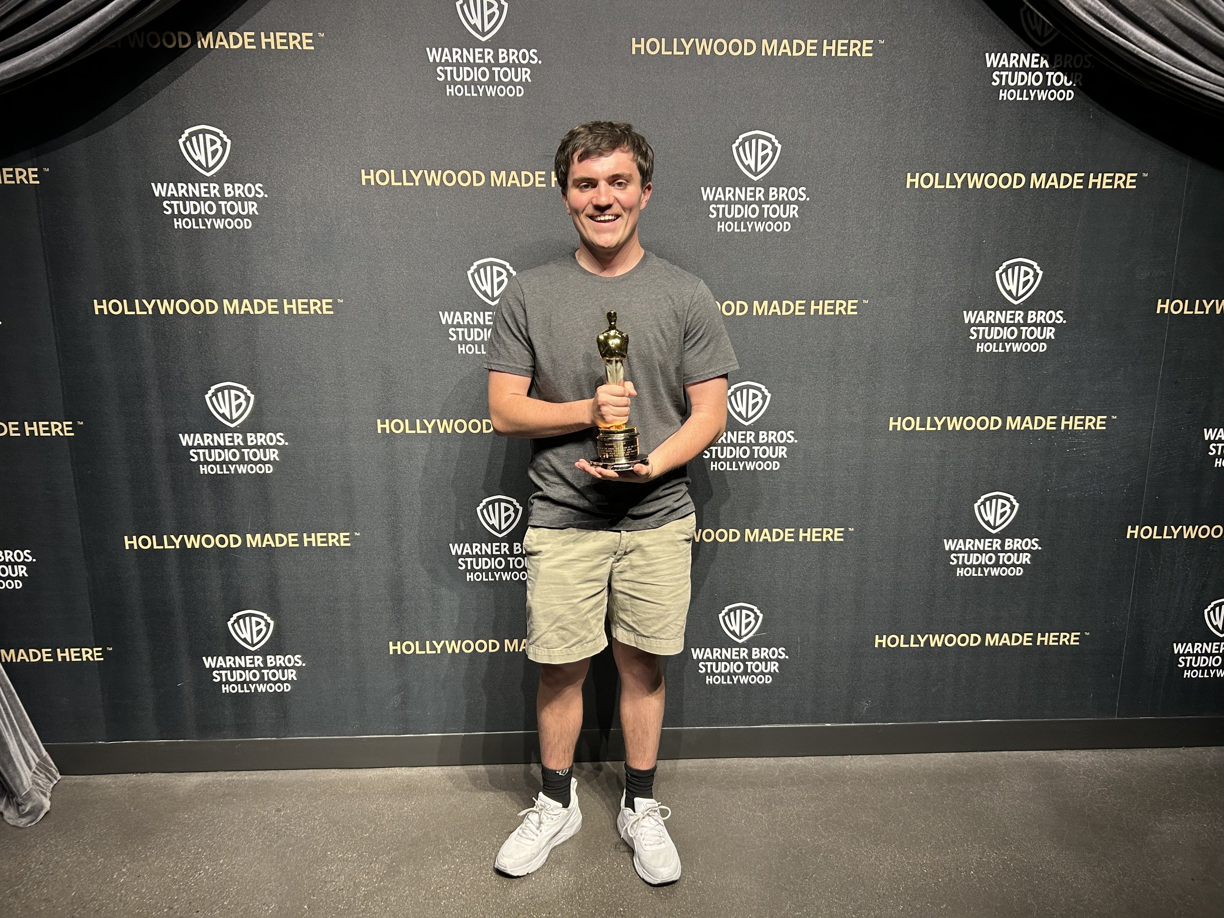 A young man smiling and holding a Tony Award trophy in front of a black backdrop with the Warner Bros. Studio Tour Hollywod logo and the text 'Hollywood Made Here' repeatedly printed on it.