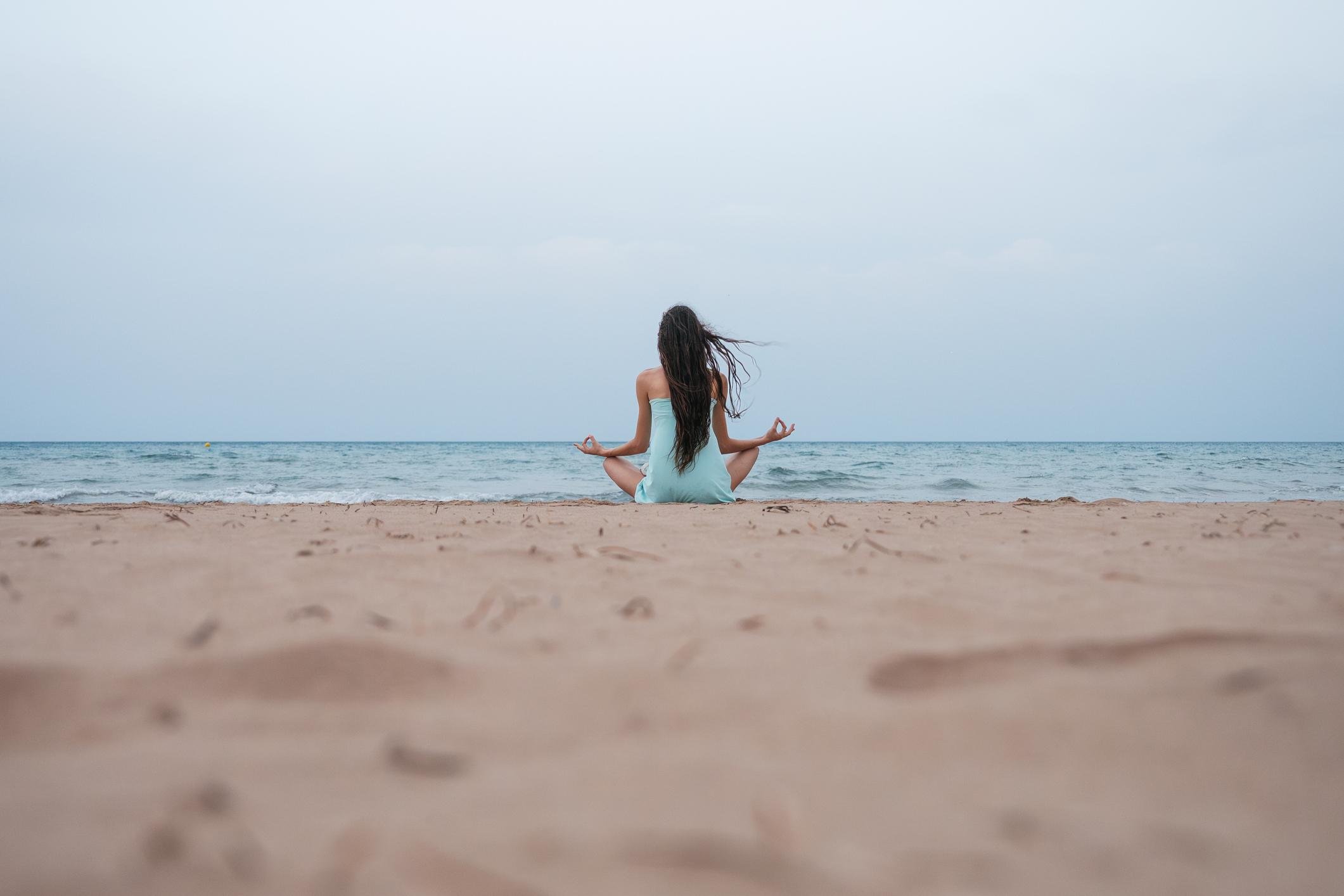 Person practicing meditation on a beach, facing the ocean under a cloudy sky