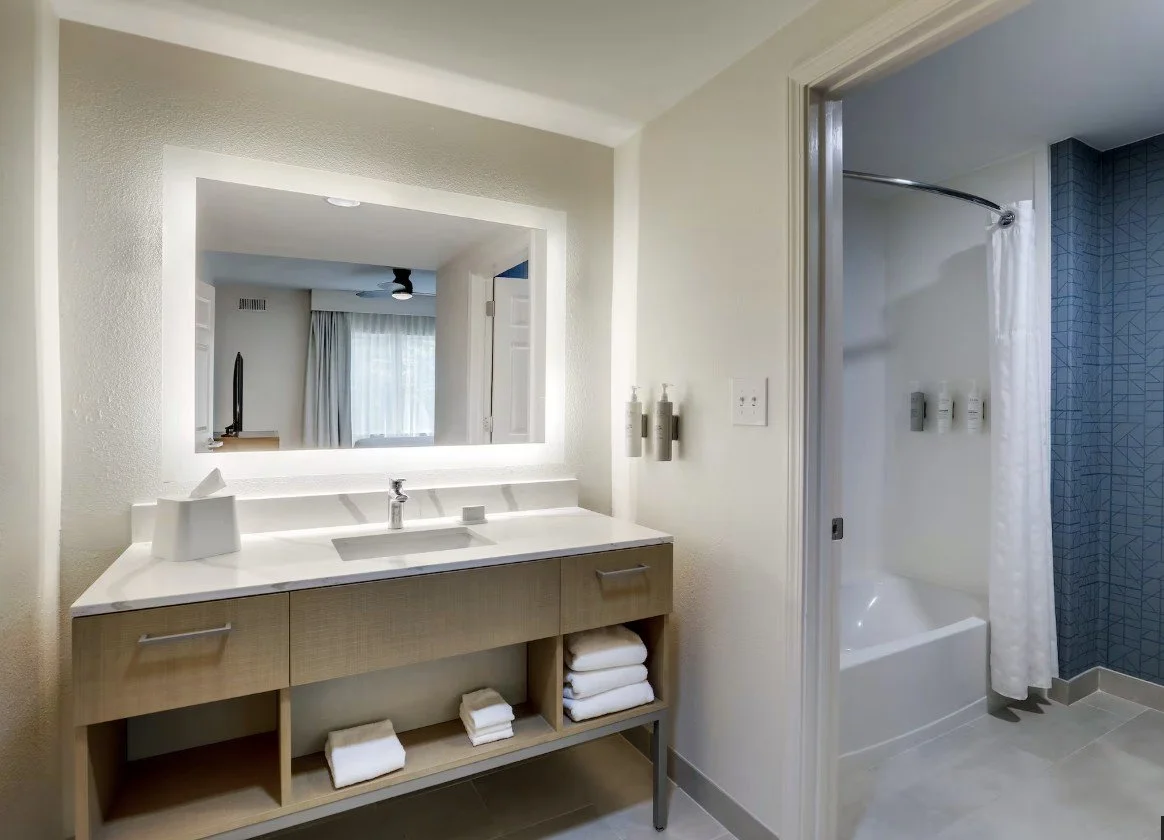 Bathroom with large mirror, sink with countertop, soap dispenser, tissue box, shelves with towels, and a separate shower area with curtain and blue-tiled wall.