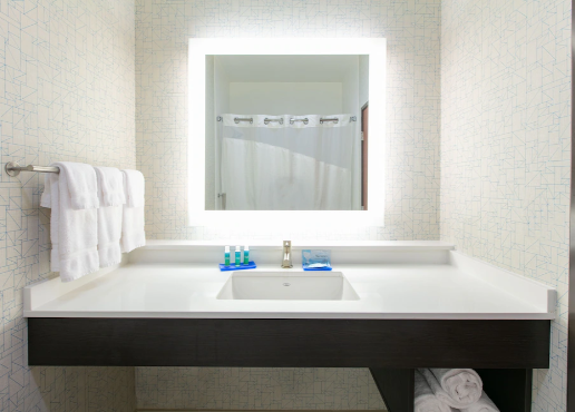 Bathroom vanity with a large illuminated mirror, a white sink, and a dark brown cabinet, with folded towels on the left and a shelf with rolled towels on the right.