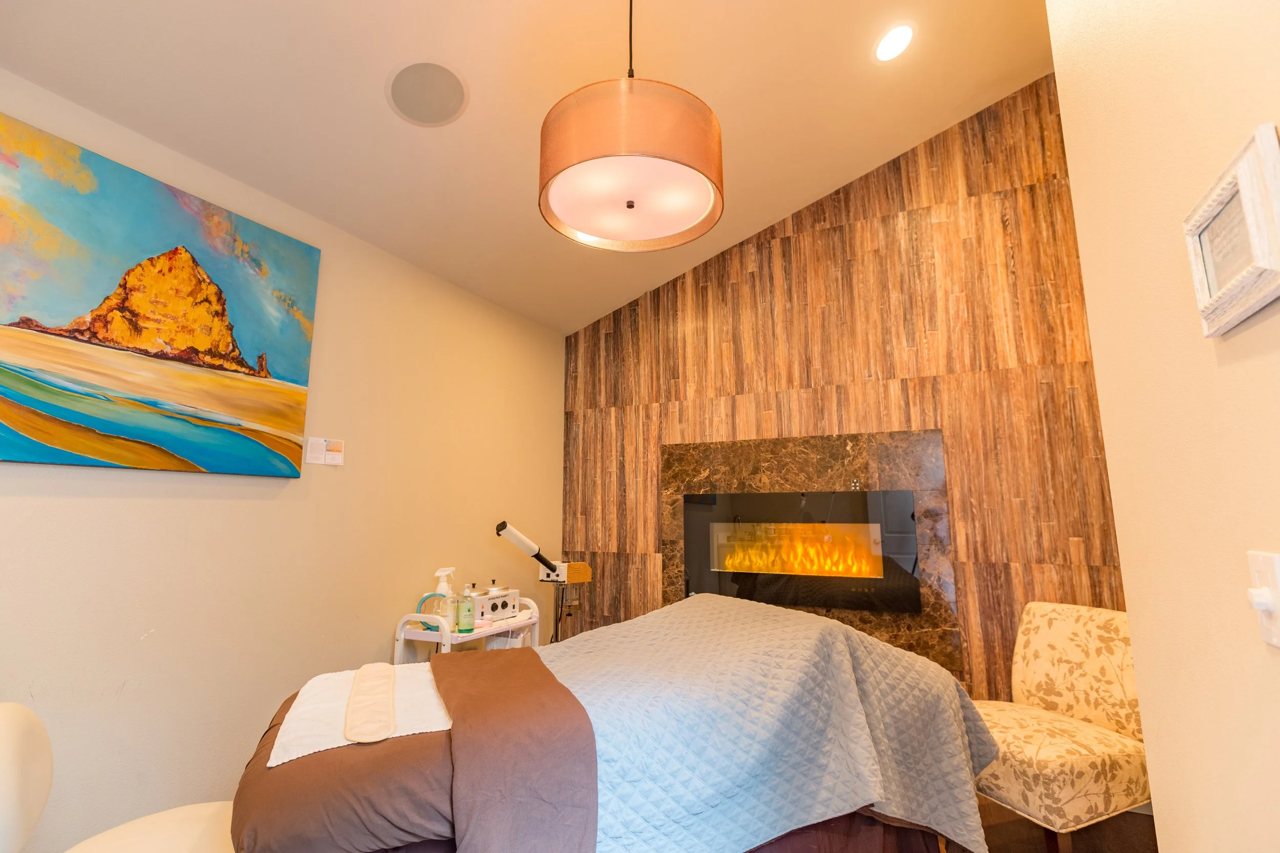 A bedroom with a bed covered in white and brown bedding, a beige chair with floral pattern, a colorful abstract painting of a mountain and ocean, wood-paneled wall, a fireplace, a hanging light fixture, and various medical or skincare supplies on a small white cart.