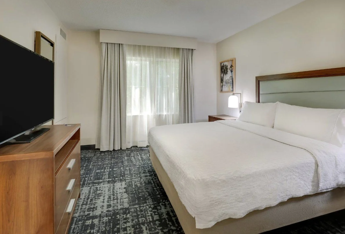 Hotel room with a large bed, white bedding, nightstand with lamp, window with curtains, artwork on the wall, flat-screen TV on wooden dresser, patterned carpet.