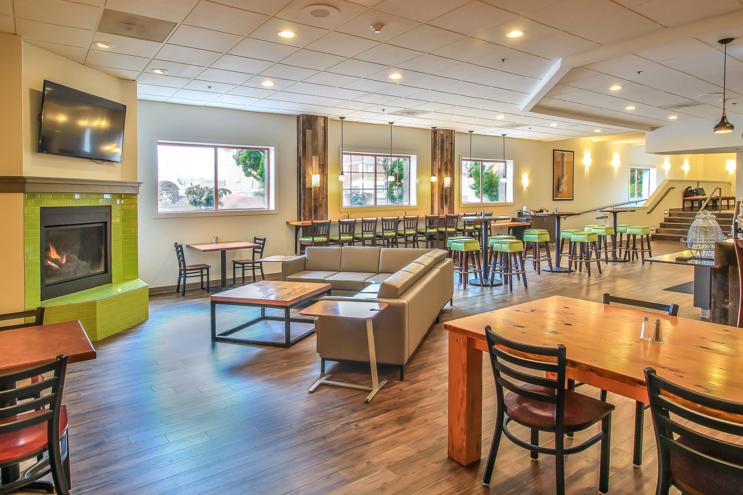 Interior view of a casual restaurant or cafe with wooden tables and chairs, a beige sofa, a green tiled fireplace, large windows, and hanging lights.