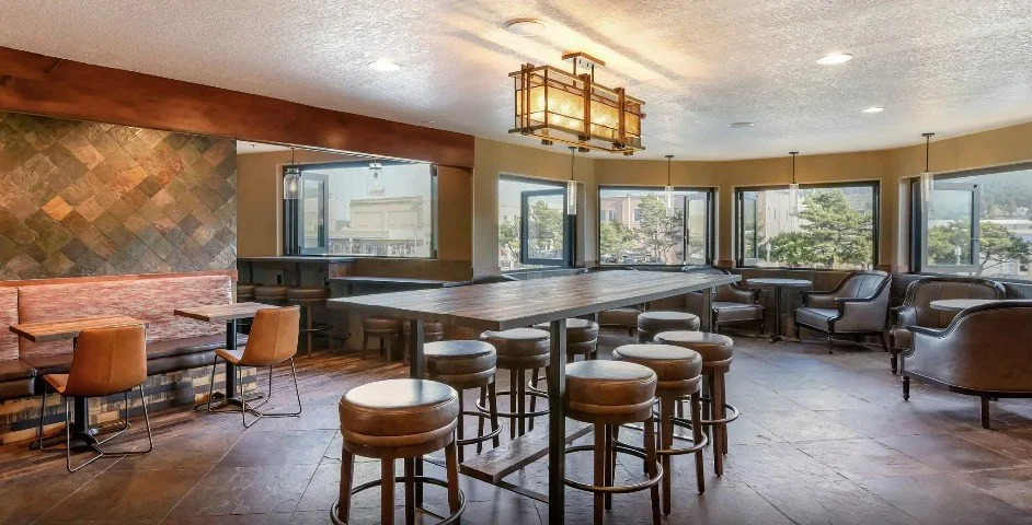 An interior view of a modern lounge or dining area with large windows, a central high-top table with stools, and comfortable seating including armchairs and a bench.