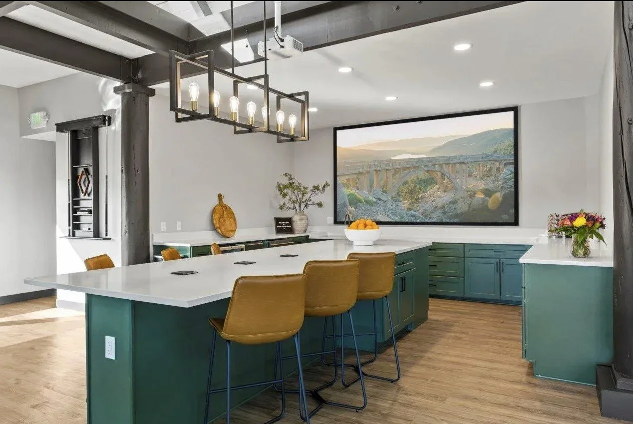 Modern kitchen in a multi-family lounge area with white countertops, green cabinets, and mustard yellow chairs around a large island.