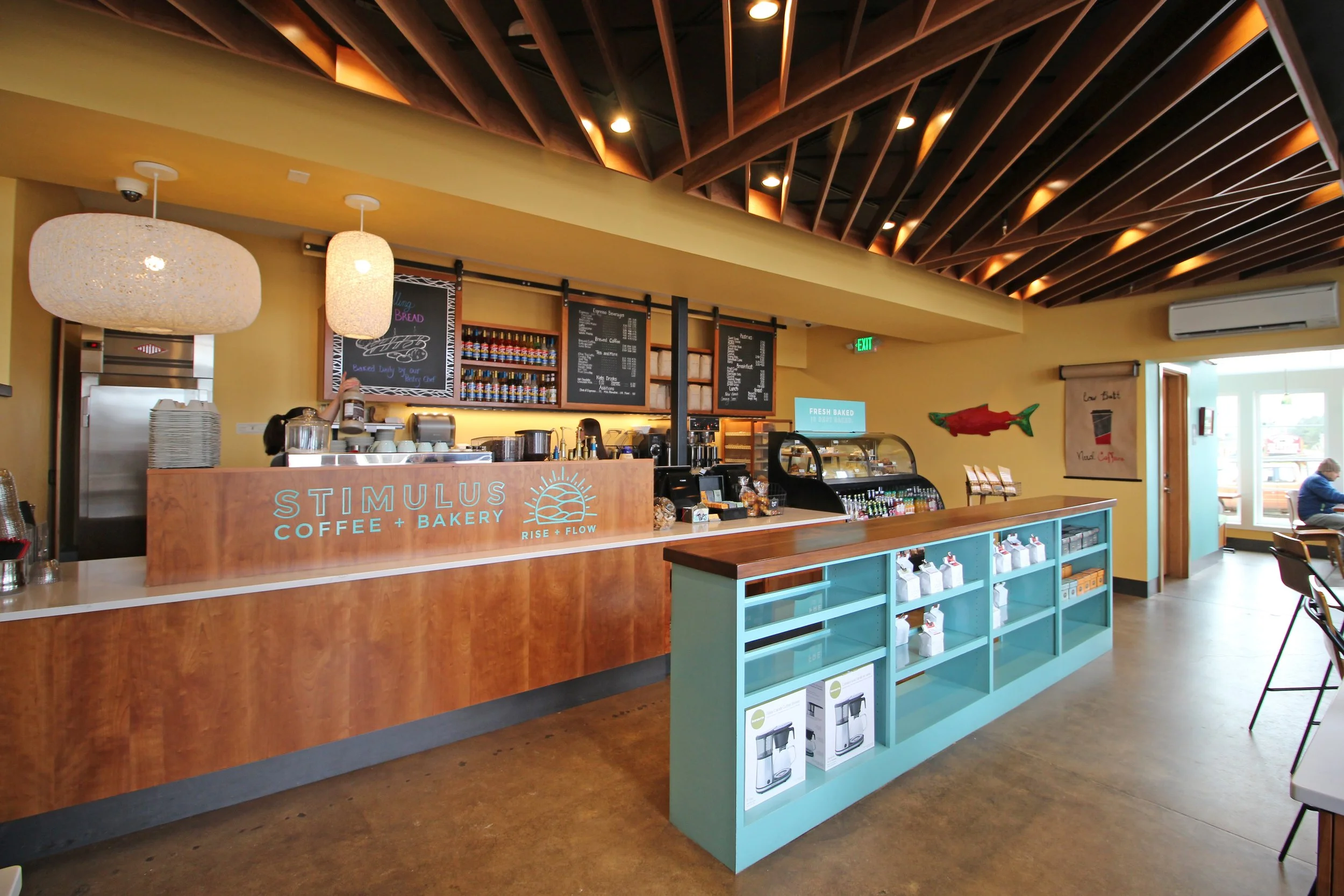 Interior of a coffee shop with a wooden counter, menu boards, hanging lamps, and a turquoise display shelf, featuring decorative fish on the wall.