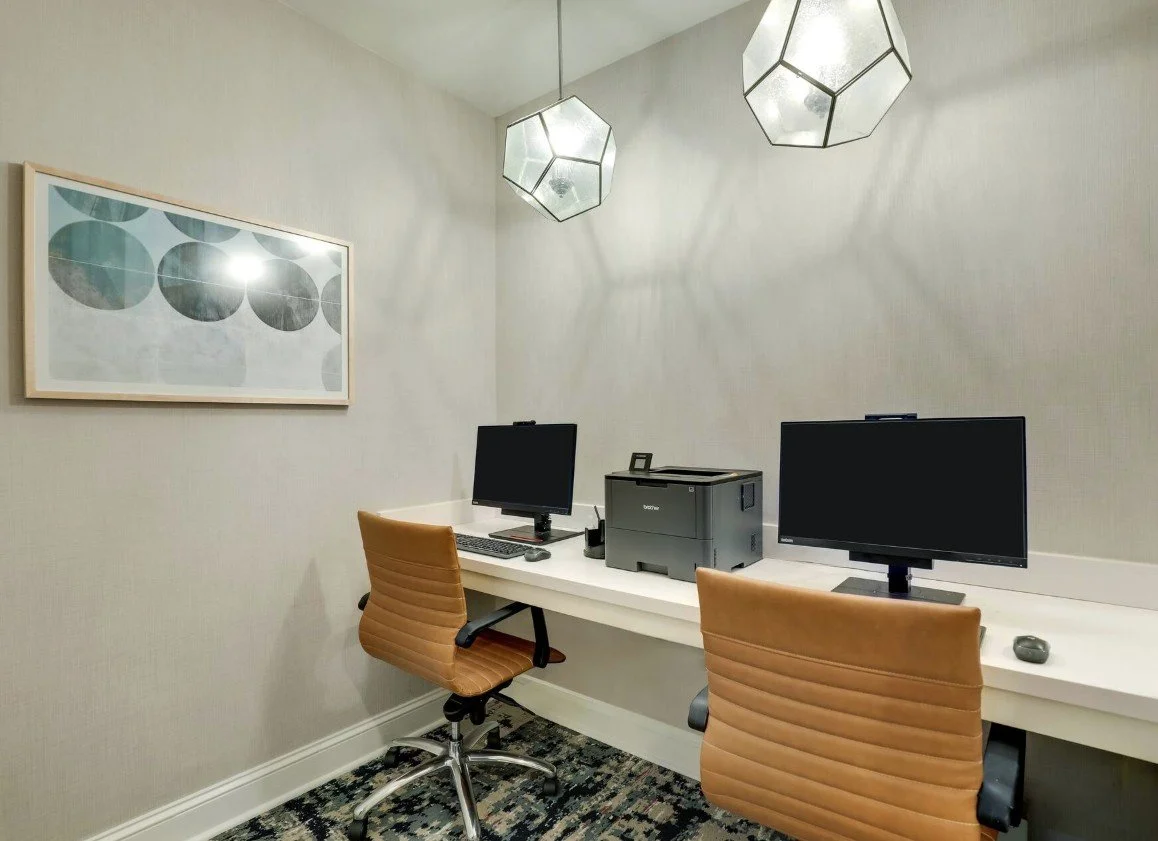 An office space with a white desk, two tan chairs, two computer monitors, a multifunction printer, and a framed geometric artwork on the wall, lit by two hexagonal pendant lights.