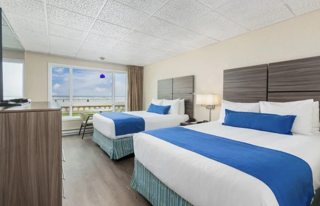 Hotel room with two double beds, blue bedding, wooden headboards, bedside lamp, window with ocean view, and a dresser.