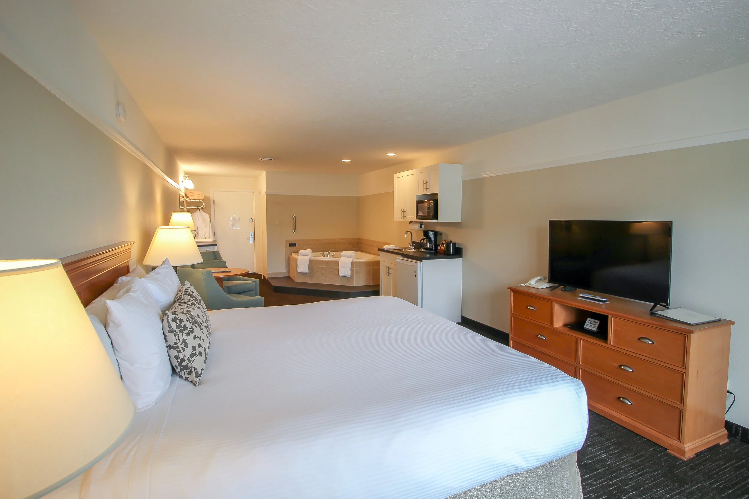 Hotel room with a bed, nightstand lamps, a sitting area with chairs, a bathroom with a bathtub, a kitchenette, and a television.