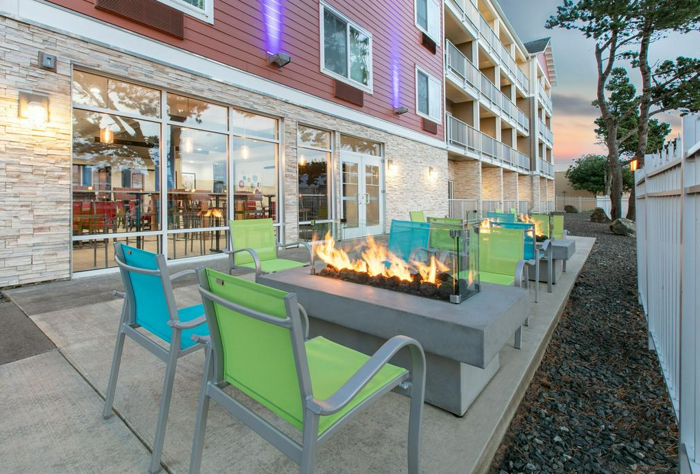 Outdoor seating area with colorful chairs around a fire pit, adjacent to a building with large glass doors and an interior dining space, with trees and a sunset sky in the background.