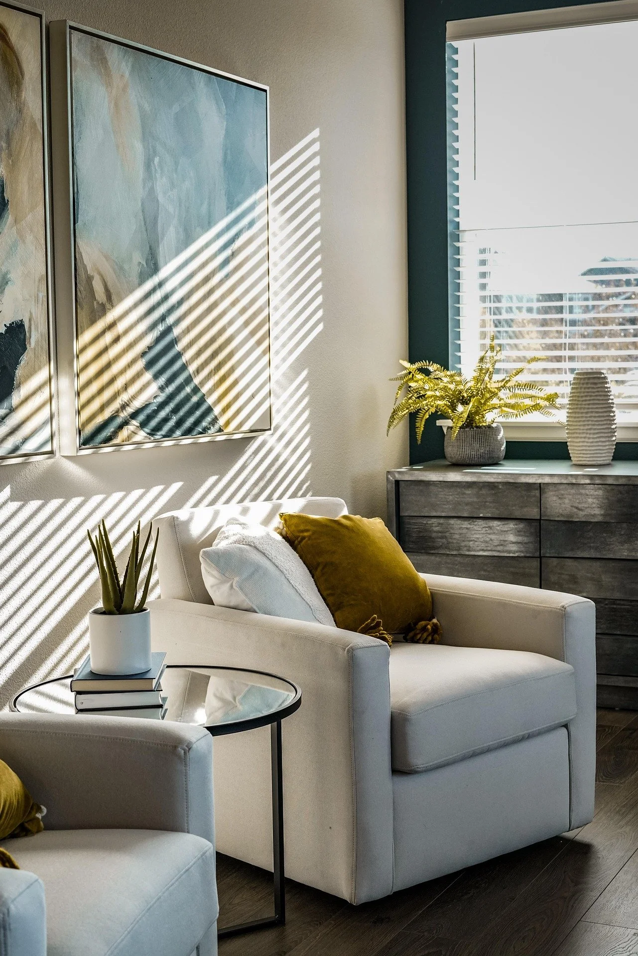 A cozy living room with a white sofa, a glass coffee table, and decorative pillows. Sunlight creates striped shadows on the wall, and a window with blinds shows an outdoor view. There are potted plants and artwork on the wall.