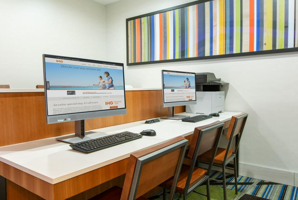 An empty office cubicle with two computer monitors, a keyboard, and a mouse on a white desk. Behind the desk is a wood-paneled wall with a colorful striped painting above.