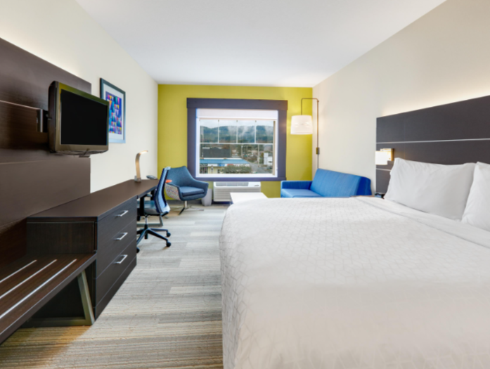 A hotel room with a large bed, a blue sofa, a desk with a chair, a mounted TV, and a window showing an outdoor view.