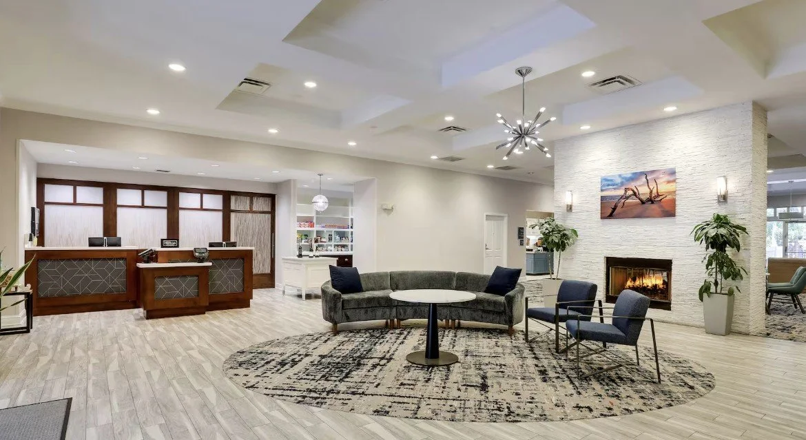 A modern hotel lobby with a reception desk, seating area with chairs and a sofa, fireplace, and artwork on the wall.