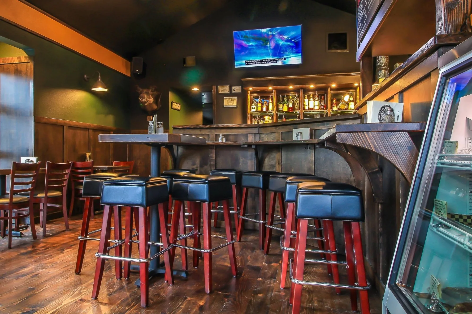 Interior of a bar or restaurant with a bar counter stocked with various liquor bottles, several black bar stools with wooden legs arranged around the counter, and a television mounted on the dark-colored wall displaying a news broadcast. There are some chairs and tables in the foreground, and a refrigerator or display case on the right side.