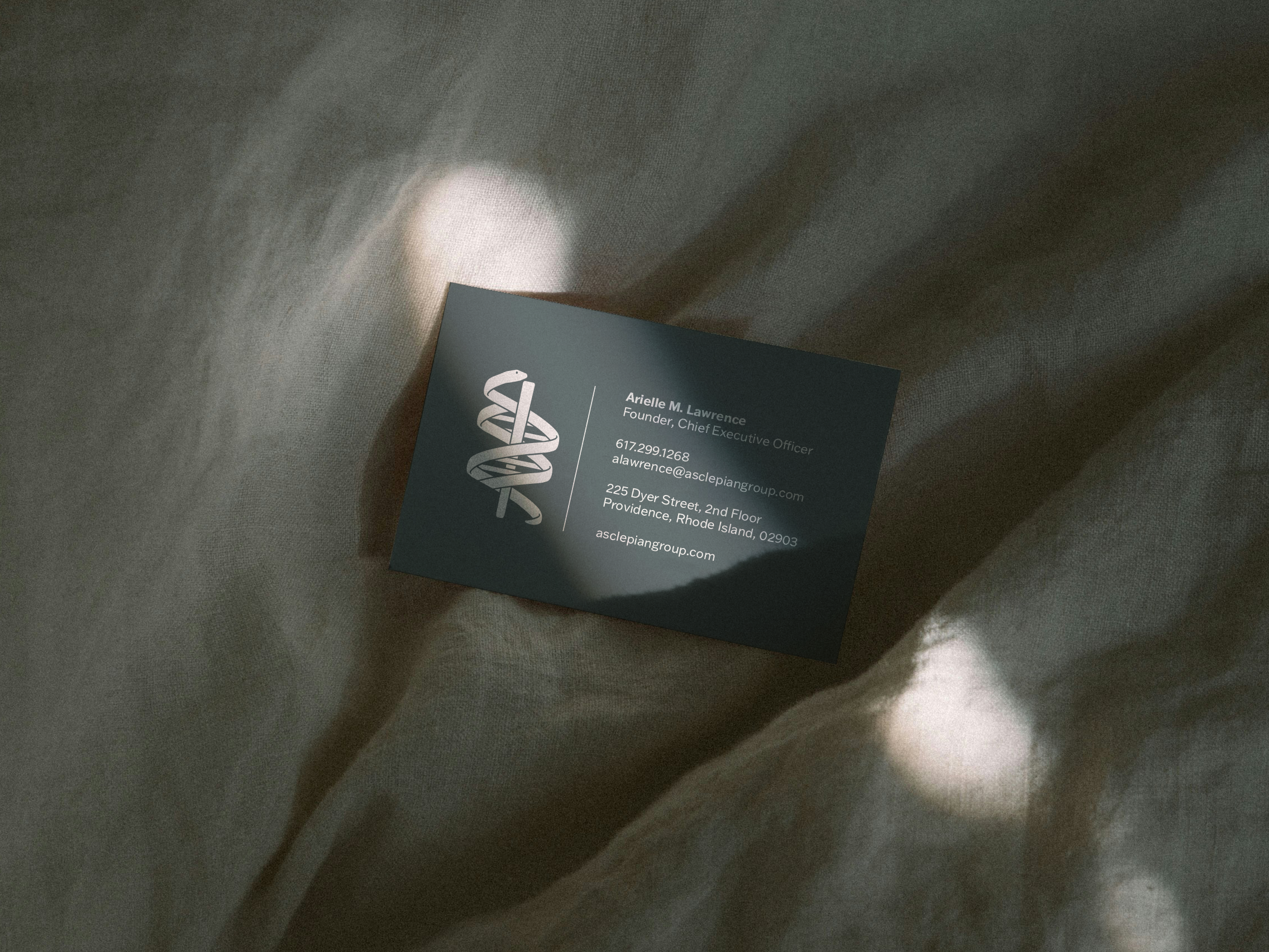 A dark-colored business card with a DNA helix logo and white text, displaying contact information, resting on wrinkled beige fabric with sunlight casting shadows.
