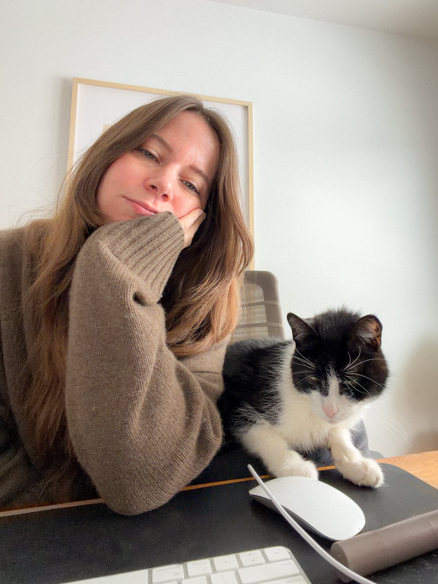 A woman with long brown hair resting her head on her hand next to a black and white cat sitting on a desk in front of a white computer mouse and keyboard, with a white wall and framed artwork in the background.