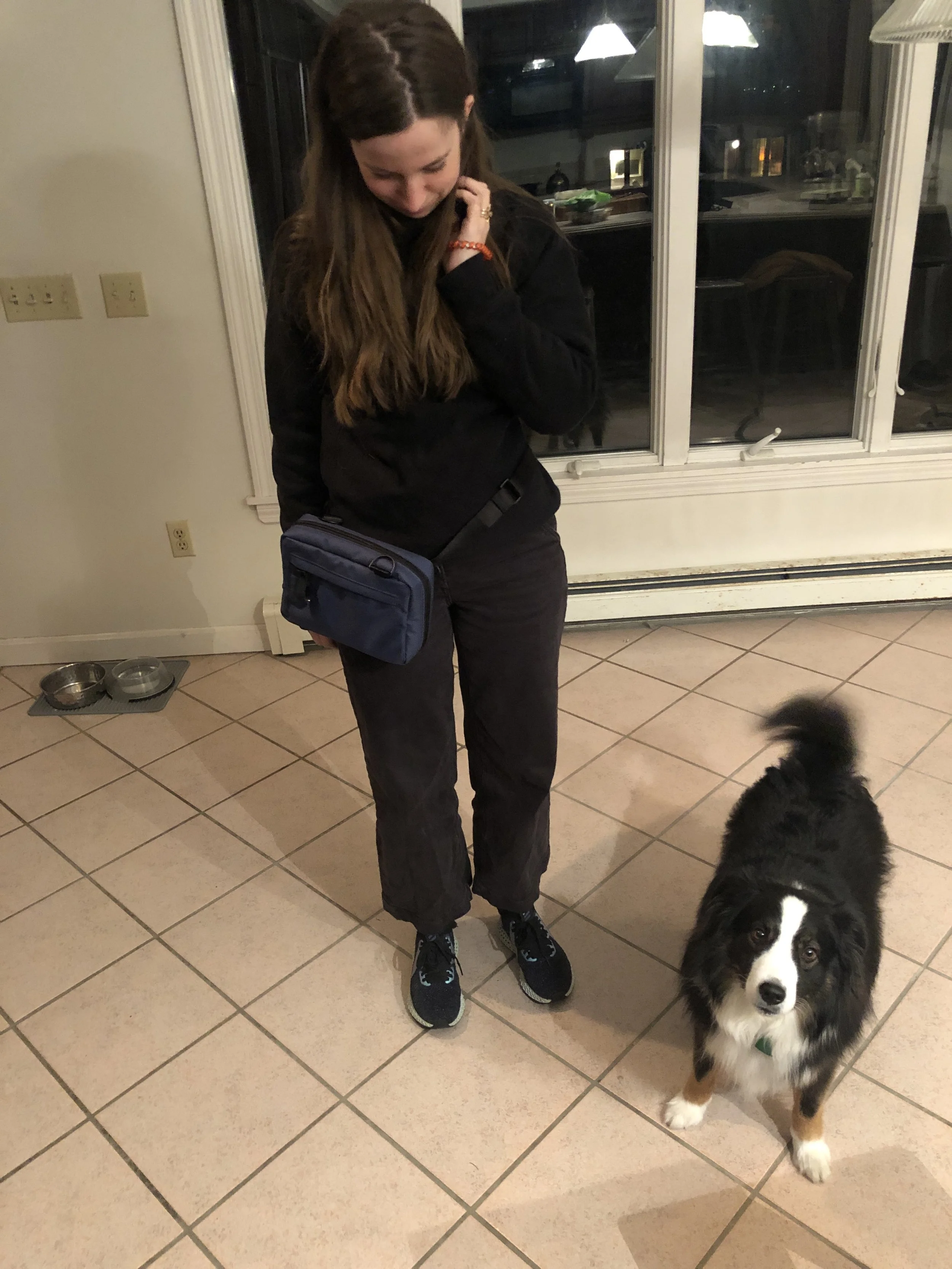 A woman with long brown hair, wearing a black jacket, black pants, and black sneakers with teal accents, stands in a room with tiled floor and large windows at night. She is looking down and touching her neck with her left hand. A border collie puppy with black, white, and brown fur stands next to her, looking directly at the camera. Behind her, there are pet food and water bowls on the floor, and a kitchen visible through the window.