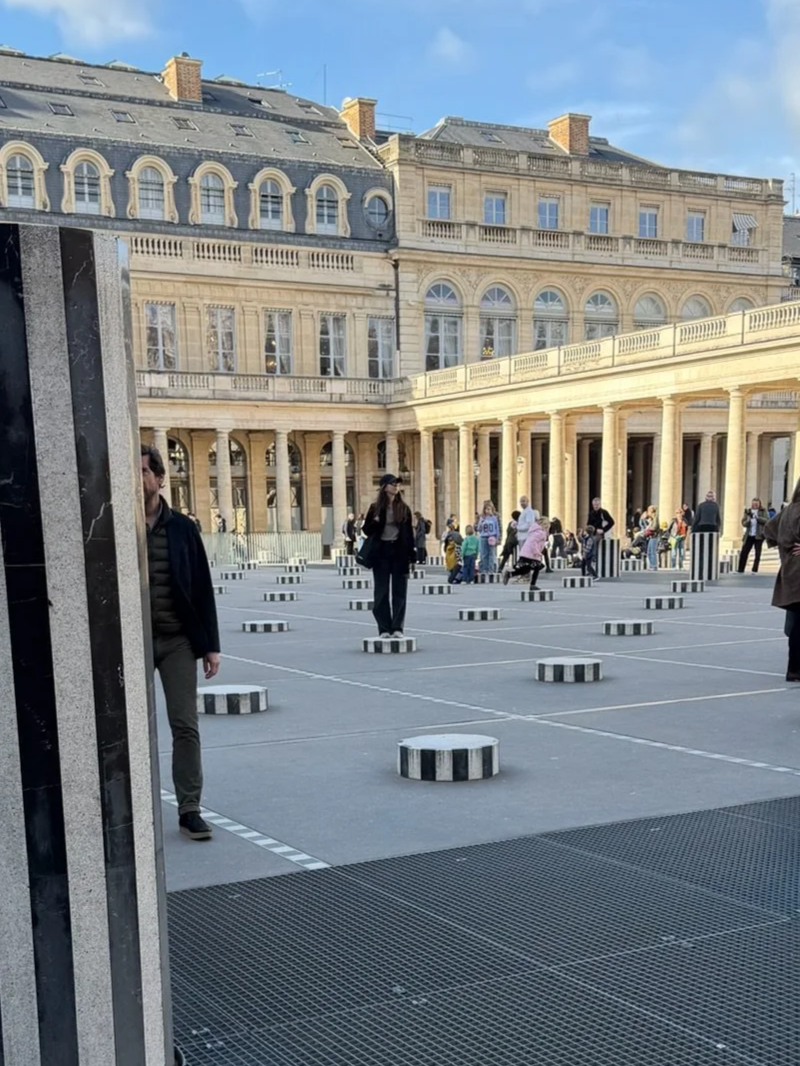 People walking and standing around a courtyard with striped cylinder-shaped seating, surrounded by historic European-style buildings with arched windows and columns, on a clear day.