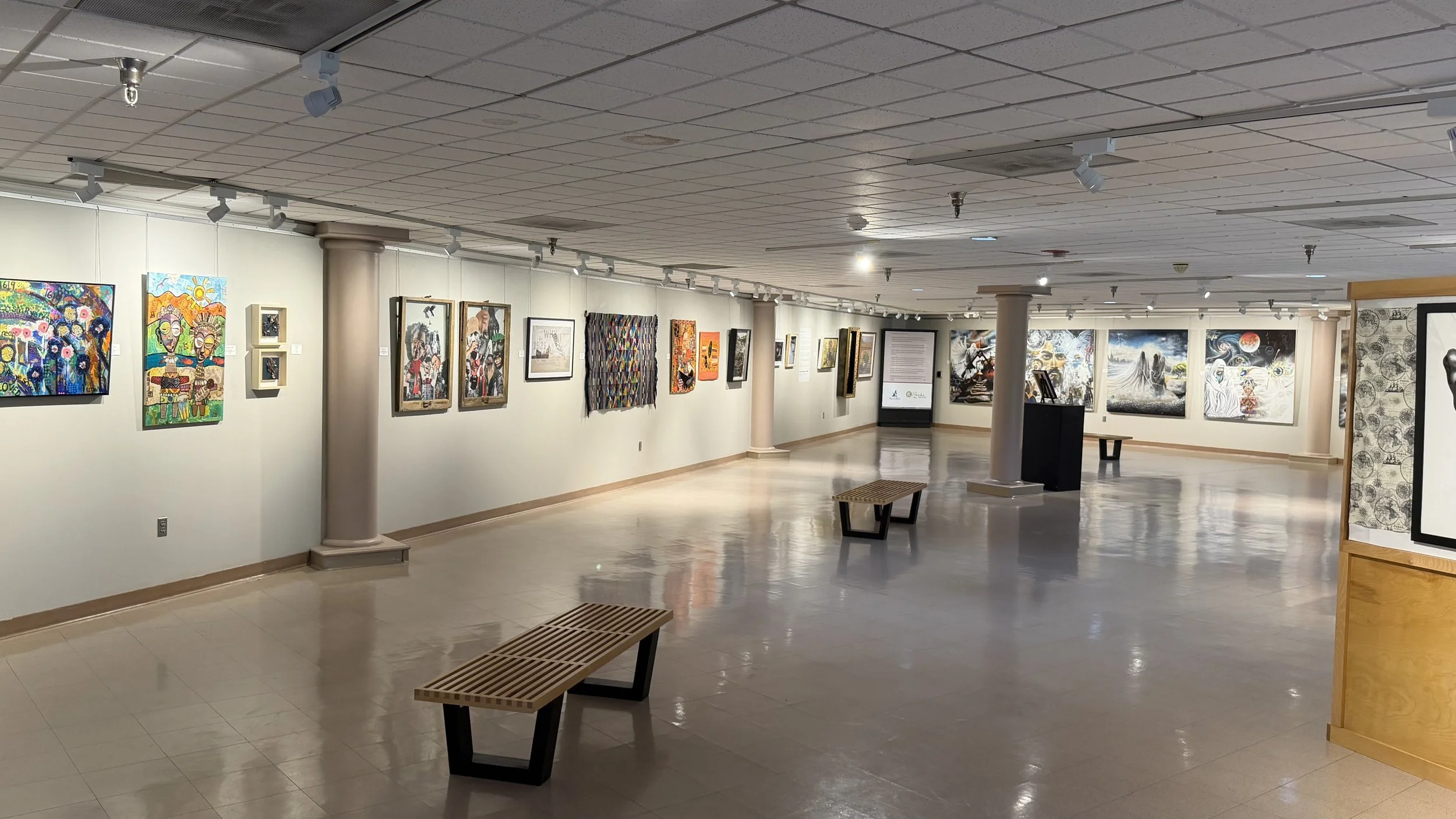 An art gallery with white walls, wall-mounted paintings, and track lighting illuminating the artwork. Main gallery at the Bruce R. Watkins Cultural Heritage Center featuring rotating artwork and exhibits along white walls