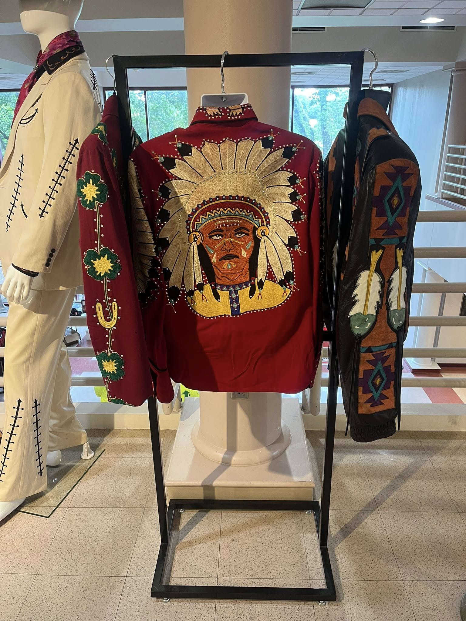 Display of western cowboy fashion with a focus on a red jacket featuring a large embroidered Native American chief with a feather headdress. Alongside, on the left, there is a white suit with black stitching details, and on the right, a black jacket 