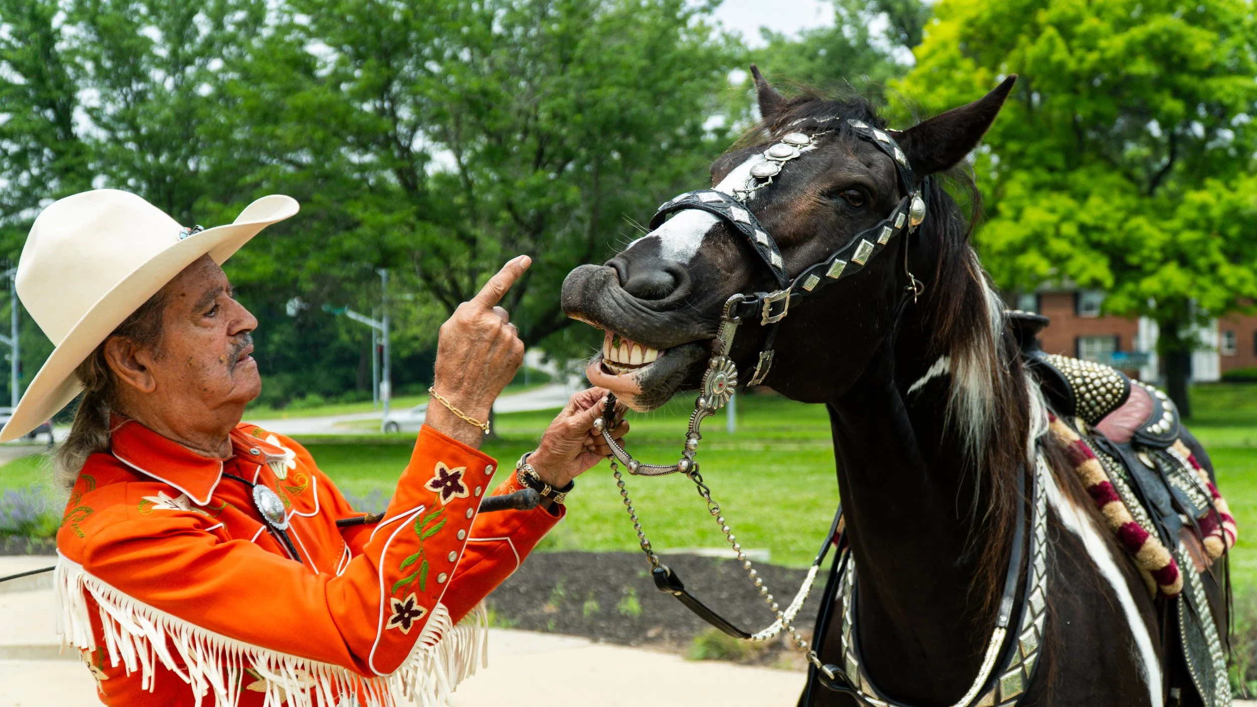 Rex Purefoy dressed in traditional clothing, including a large white hat and orange fringed shirt, gently touching the nose of a decorated black horse with a white blaze on its face, set against a background of green trees.