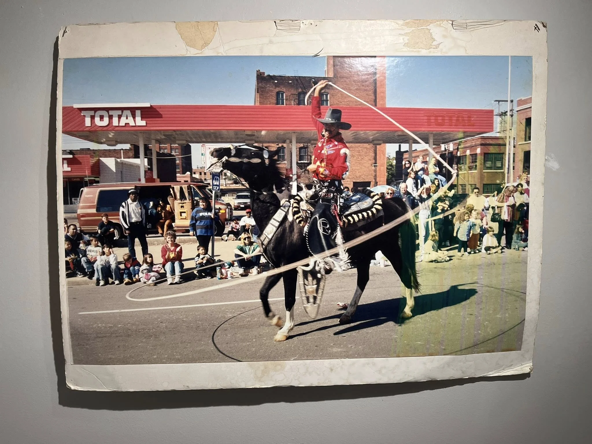 A street parade scene with Rex Purefoy dressed as a cowboy riding a horse, surrounded by a crowd of children and adults. The background features a red and white gas station and brick buildings.