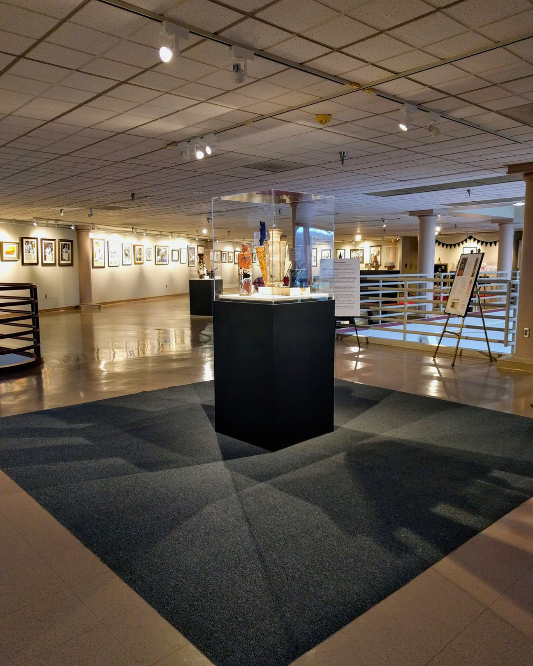 An art gallery with framed artworks on the walls, a display case with sculptures, and informational signs on stands.