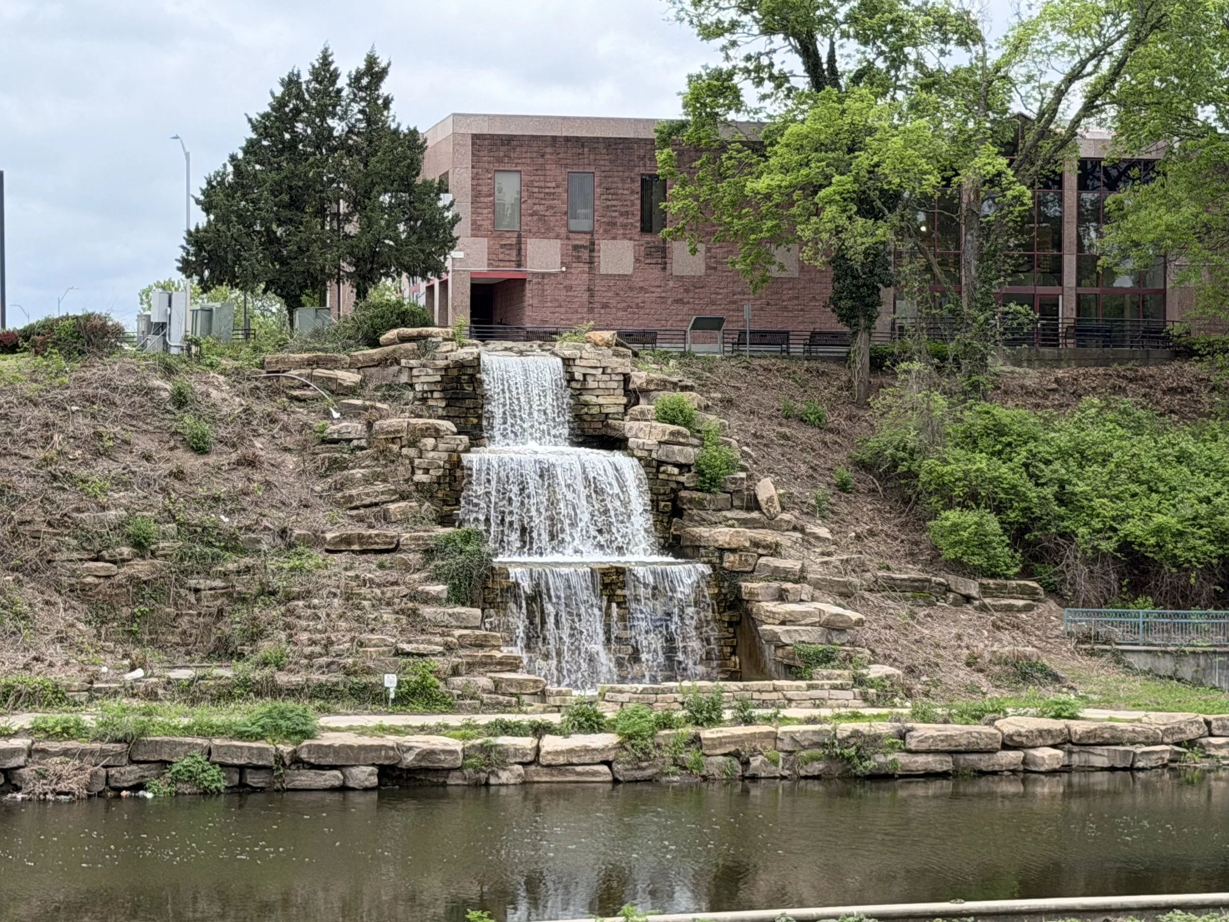 A water feature with a small waterfall cascading over stacked rocks, situated in front of a modern brick building with windows. Green trees and a body of water are in the foreground.