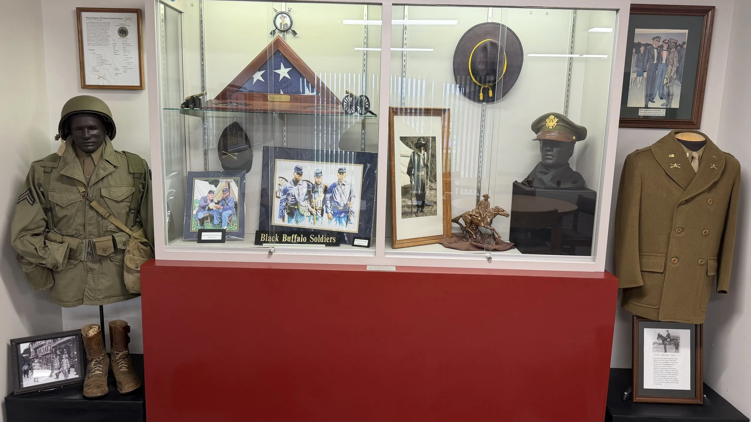 Display case featuring military memorabilia including mannequins dressed in military uniforms, framed photos, a miniature horse sculpture, medals, a black and gold framed display labeled "Black Buffalo Soldiers," and various framed documents and phot