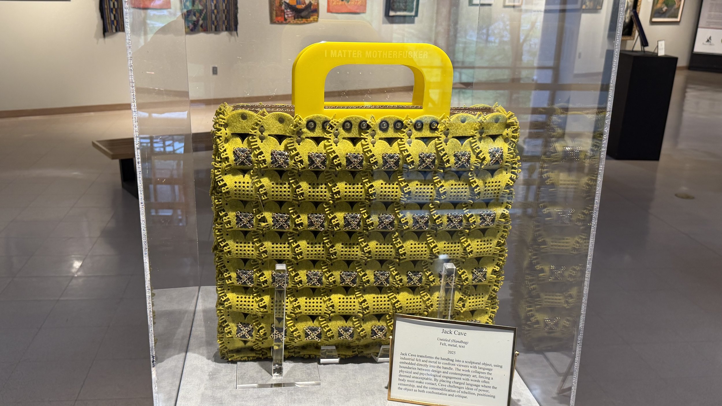 A yellow handbag-shaped sculpture with the words "I MATTER MOTHERFUCKER" on the handle, featuring various patterns and embellishments, displayed in a museum or gallery setting.